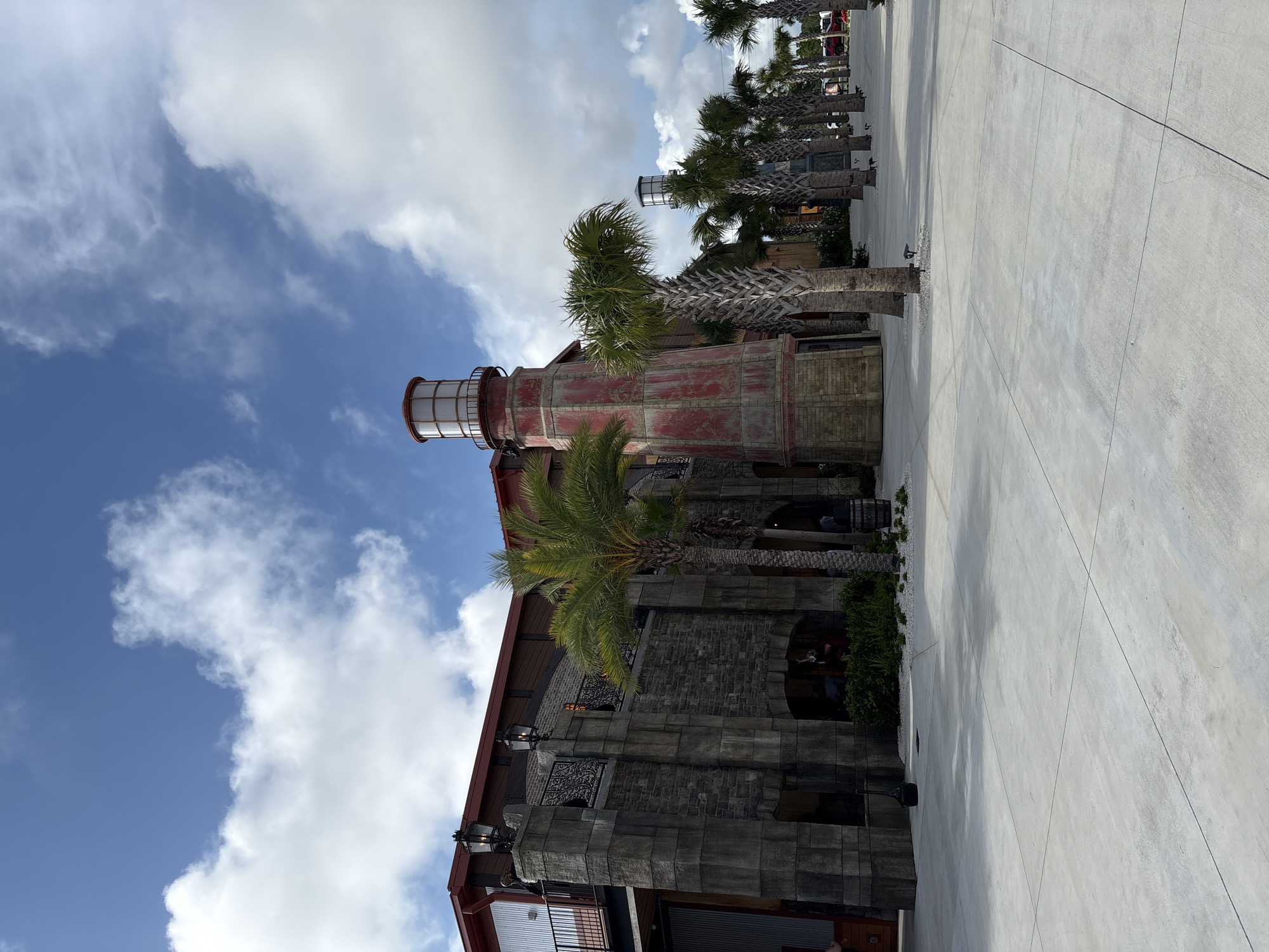 This image shows an exterior shot of a building with a distinct lighthouse-like structure on top, located in Panama City Beach, USA. The building's lower section appears to be constructed of stone or stone-like material, possibly brick, with what looks like decorative ironwork on the facade. Above that is the lighthouse tower, painted red, capped with a lantern-like structure with what may be glass panels. Palm trees are visible on both sides of the building. A concrete walkway extends to the right, suggesting a public space. The sky is blue with scattered white clouds.