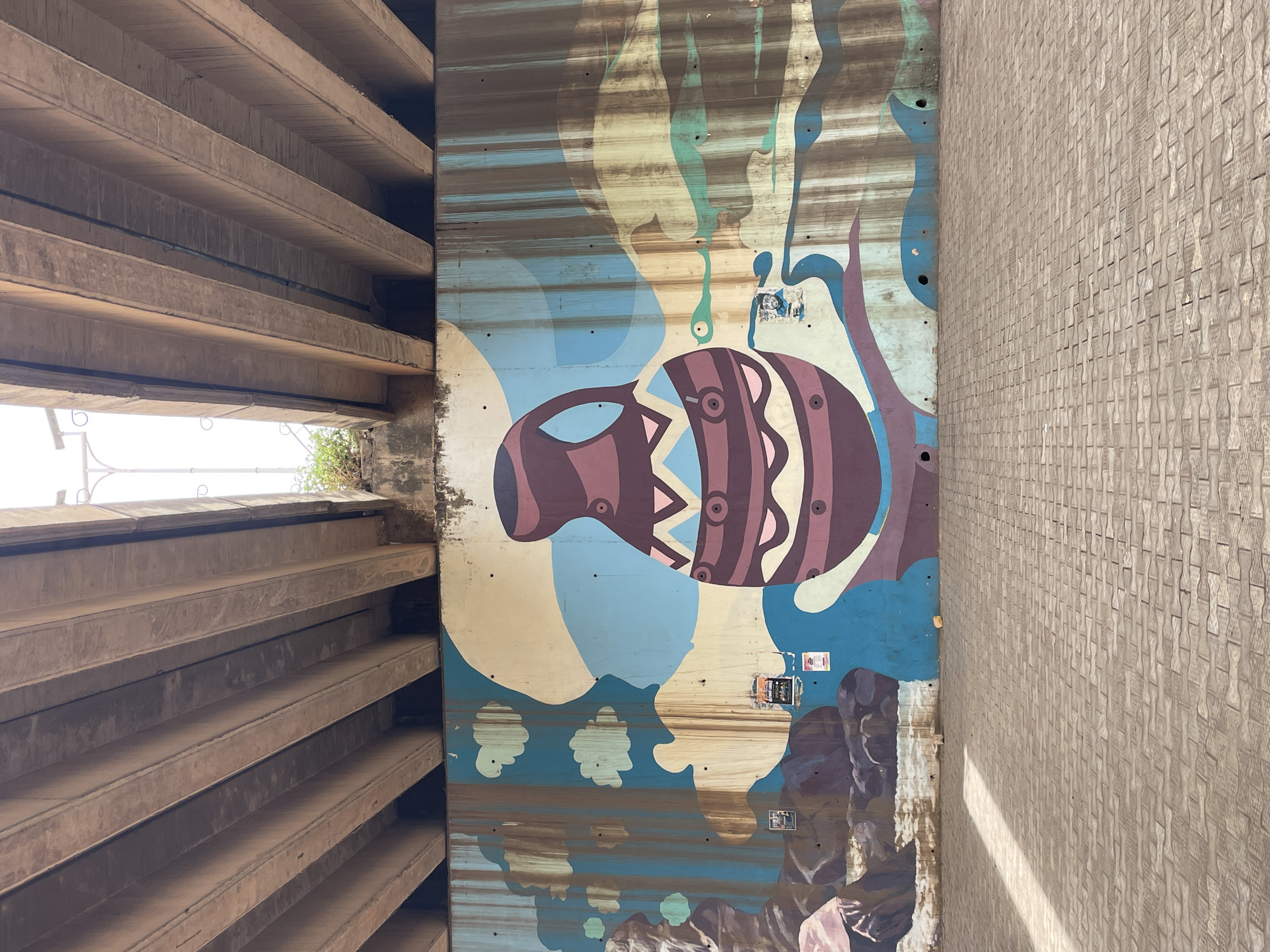 An upward, eye-level shot depicts a concrete underpass in Jos, Nigeria. On the left is a section of stacked, horizontal concrete supports. On the right, a textured, tan wall is visible. The central area features an abstract mural painted on the concrete wall of the underpass. The mural includes a brown and pink pot-like shape with geometric patterns, against a background of blue, beige, and teal colors. The bottom of the mural depicts brownish shapes resembling rocks or figures. The mural appears weathered with dirt streaks and peeling. Small stickers are affixed to the wall near the mural. Light peeks through the gaps between the concrete supports on the left.