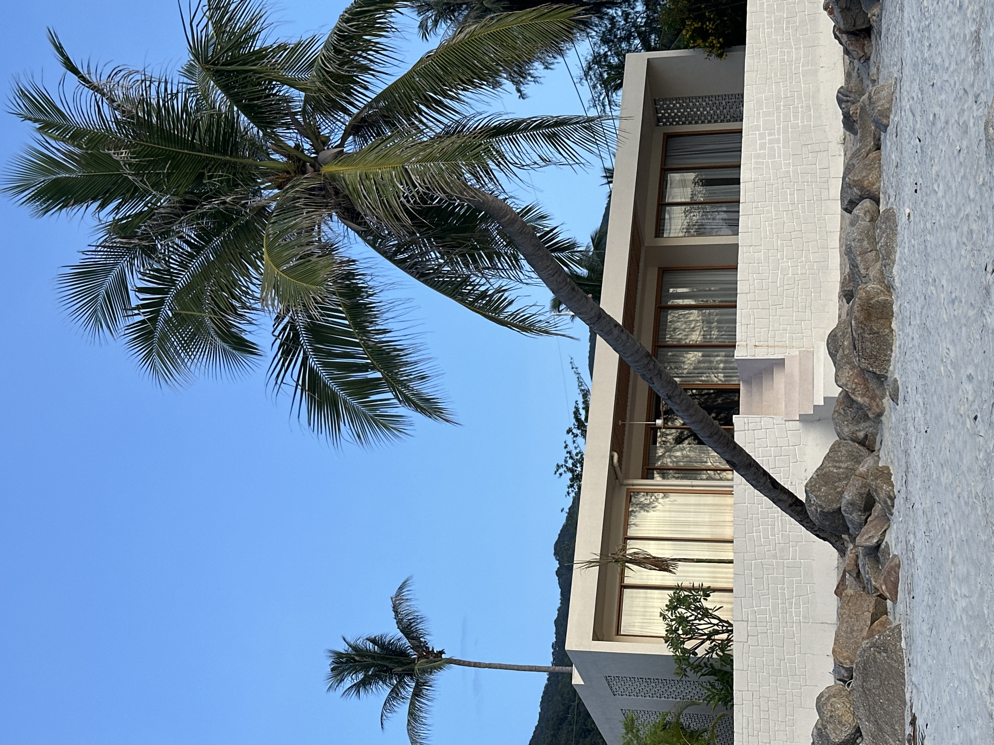 The image depicts a modern house situated on a beach in Tambon Ko Pha Ngan, Thailand. The house is a single-story structure with a light-colored exterior, featuring large windows with wooden frames and sheer curtains. A low, textured white wall with a brick-like pattern runs along the front of the property, separating it from the beach. Large rocks are scattered on the sandy beach in front of the wall. A prominent palm tree leans over the house from the right side of the frame, its fronds extending over the roof. Another palm tree stands tall in the background to the left of the house. The sky is a clear, light blue, suggesting it is daytime, possibly late afternoon given the lighting. The overall scene conveys a tropical, coastal ambiance. There are no people visible in the image.