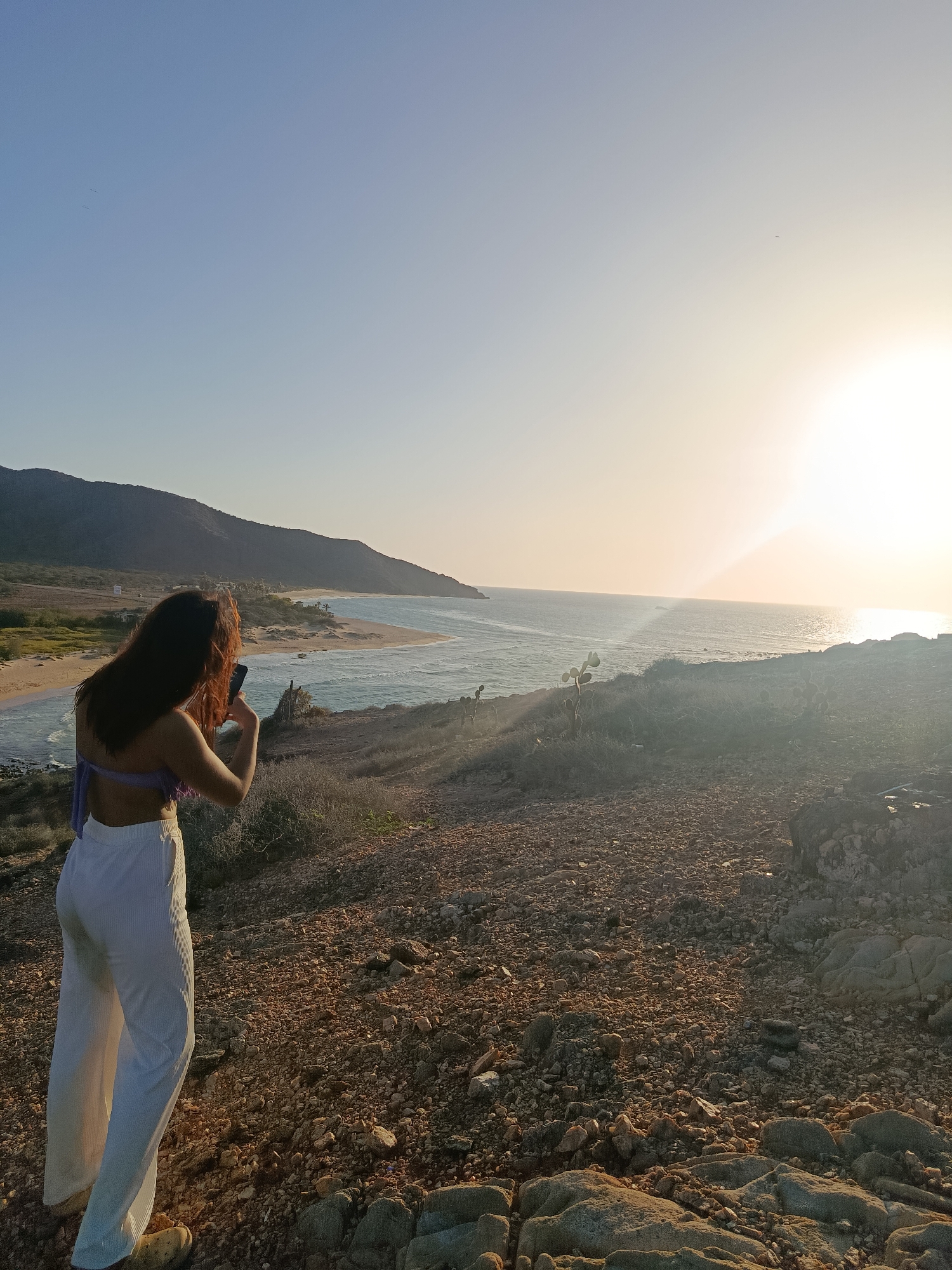 A woman stands on a rocky, arid hillside overlooking a tranquil bay. She is facing away from the camera, holding a smartphone in her hands as if to take a photo. She wears a purple halter top and flowing white pants. The bay, with its calm, blue waters, is framed by a sandy beach and a verdant, hilly landscape in the distance. The sun is setting over the horizon, casting a warm, golden glow across the sky and sea. The scene suggests a peaceful, natural environment, possibly in the vicinity of Altagracia, Venezuela, known for its coastal beauty.
