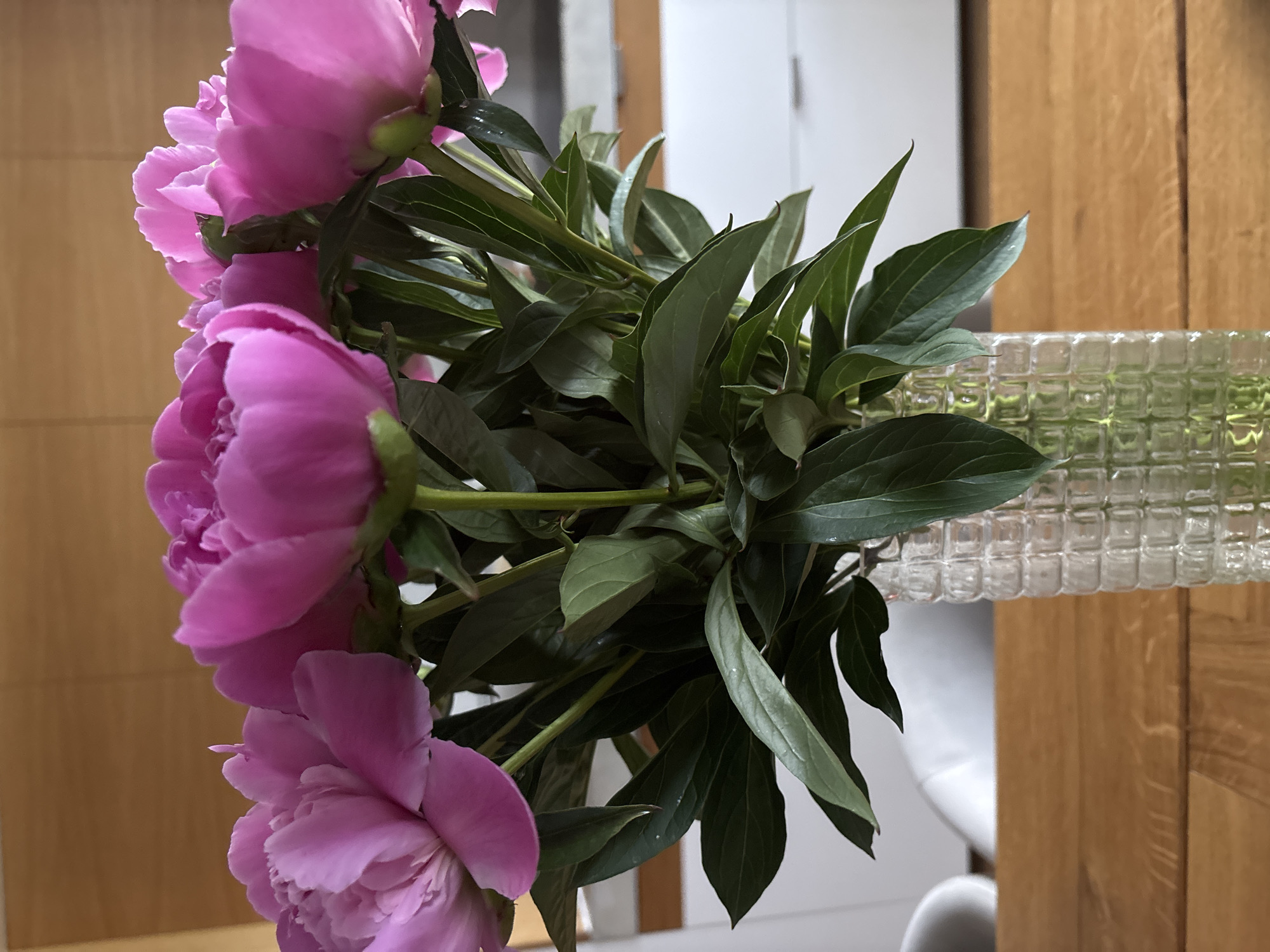 A close-up shows a bouquet of pink peonies arranged in a textured glass vase. The flowers are in full bloom, with several layers of petals. The leaves are dark green and glossy. The vase is cylindrical and made of clear glass with a grid-like pattern. The background consists of a light brown wooden surface and blurred white objects. The location is not discernable from this image.