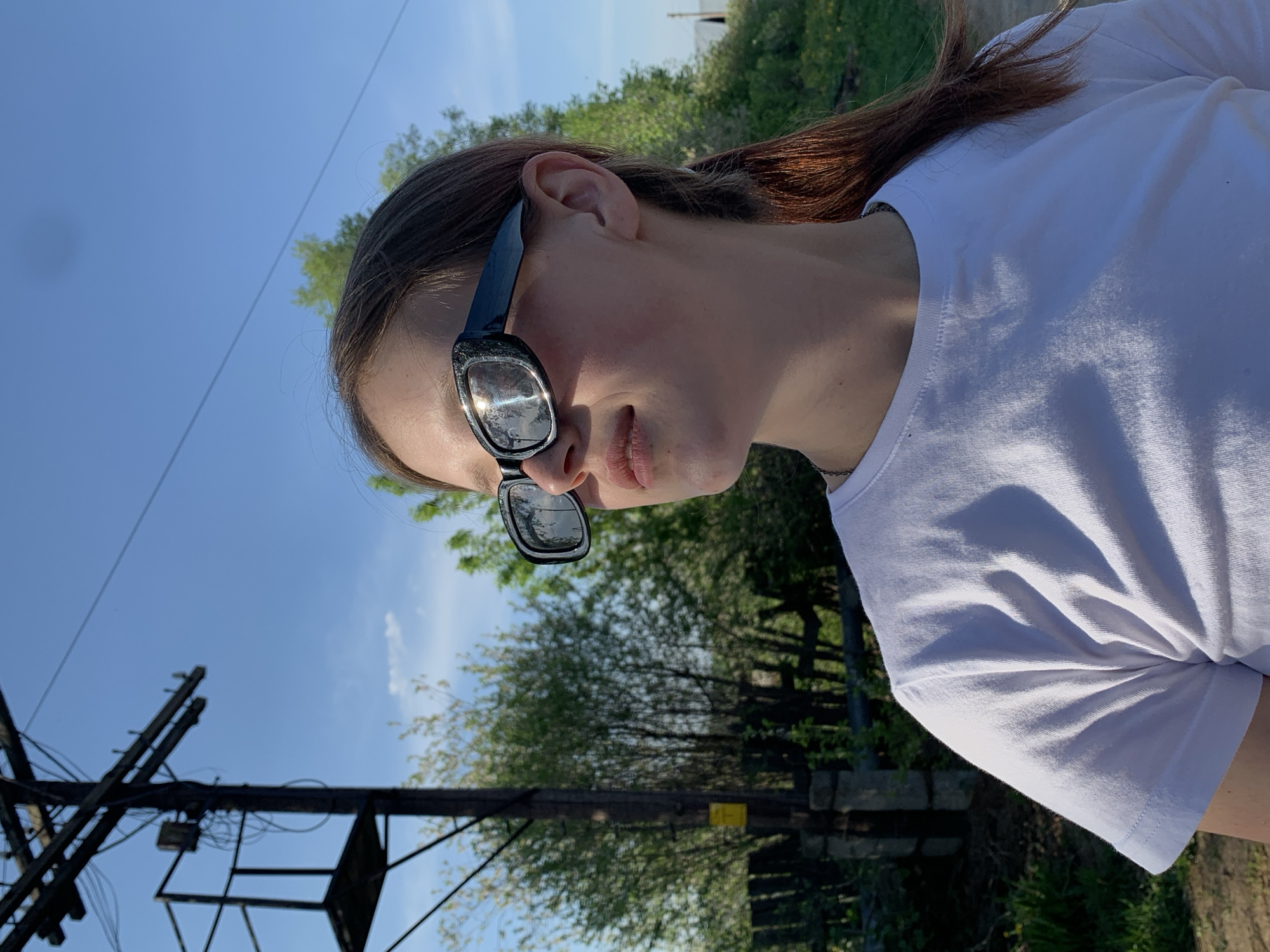 The image shows a person wearing sunglasses and a white shirt. The sunglasses reflect the sky and trees. The person is outdoors, with trees and a blue sky in the background. There is a utility pole and some vegetation behind the person, though it is difficult to confirm location in Фоки, Russia, from the image.
