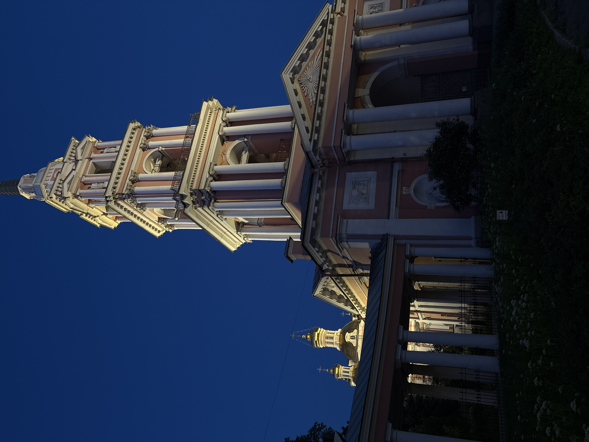 The image shows the St. Nicholas Naval Cathedral in St. Petersburg, Russia, at dusk. The cathedral's bell tower, painted in shades of pink and white with gold accents, dominates the right side of the frame. Its tall, multi-tiered structure is adorned with columns and decorative elements. To the left, the golden domes of the main cathedral rise against the deep blue twilight sky. The foreground features a dark, grassy area with sparse white flowers, and a black metal fence runs along the base of the cathedral's covered walkway. The building is illuminated, casting warm light on its architectural details. The sky is clear and dark blue, indicating nighttime. There are no people visible in the scene.