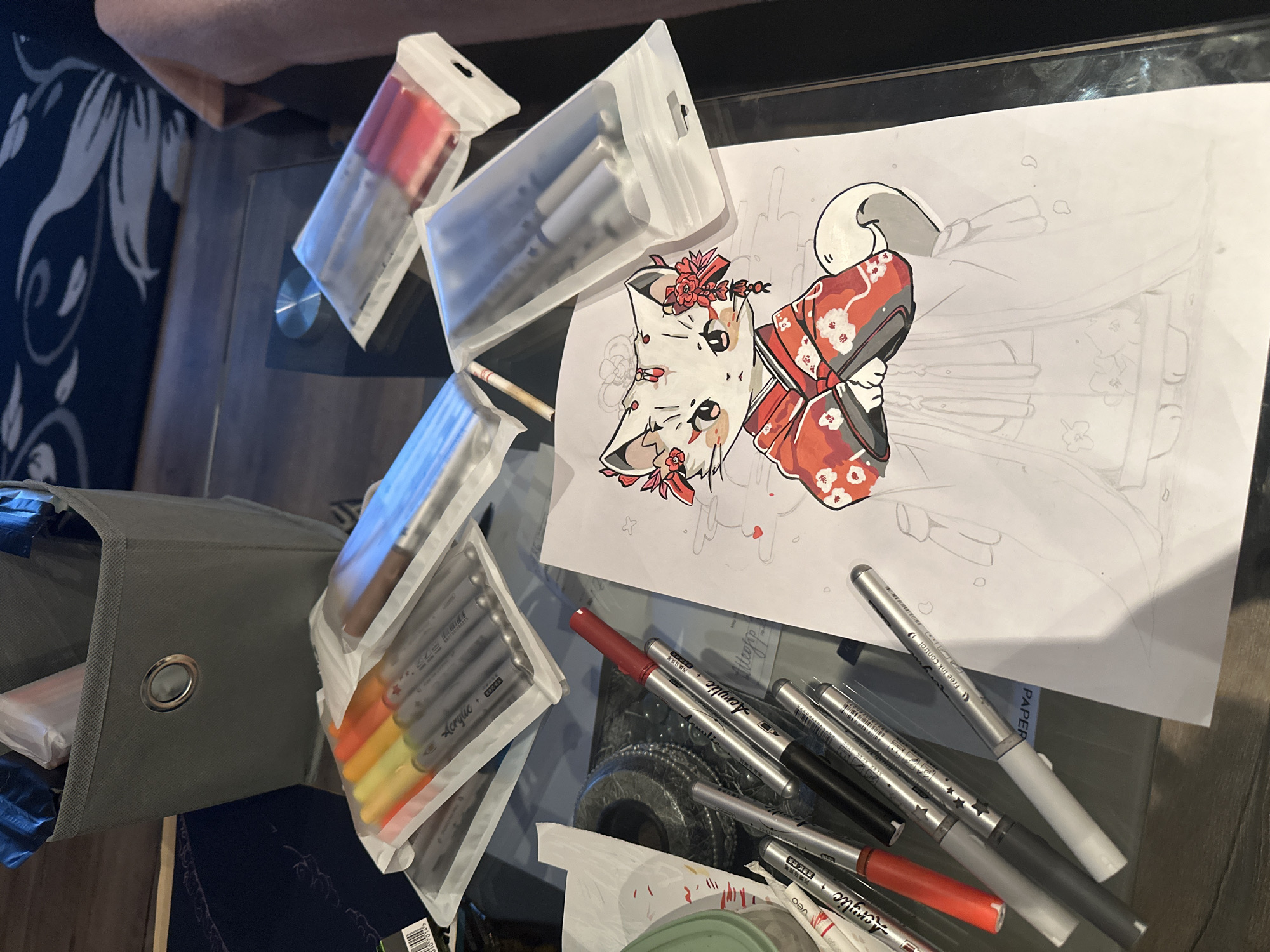 The image shows a drawing and art supplies on a glass table. The drawing depicts a stylized anime-style fox in a traditional kimono. The fox has red flowers on its head and neck. The kimono is red with white flower patterns. The rest of the drawing is a light sketch. Surrounding the drawing are several packages of markers in various color families (reds, grays, and warm tones), and loose markers are scattered near the drawing. There is a gray canvas storage cube on the left edge of the image. The location is indiscernible.