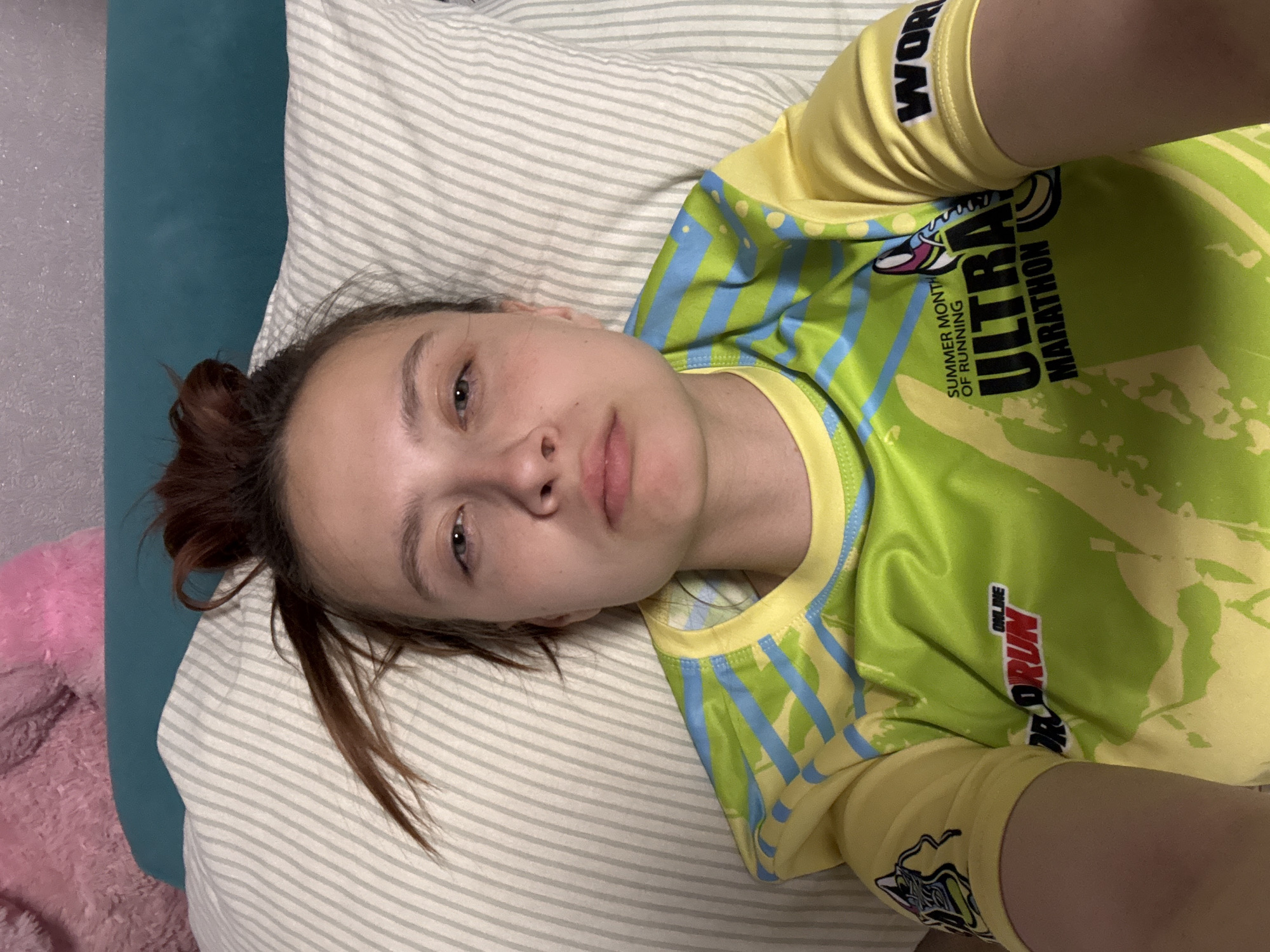 A young woman is lying on her side on a striped pillow and teal-colored bedding. She is wearing a yellow and green athletic shirt with blue stripes and various logos and text, including "Ultra Marathon" and "Pro Run." Her hair is pulled back in a messy bun with loose strands. Her face is visible with freckles and a neutral expression. A pink plush object is partially visible on the left side of the frame. The location is Прикамский, Russia.