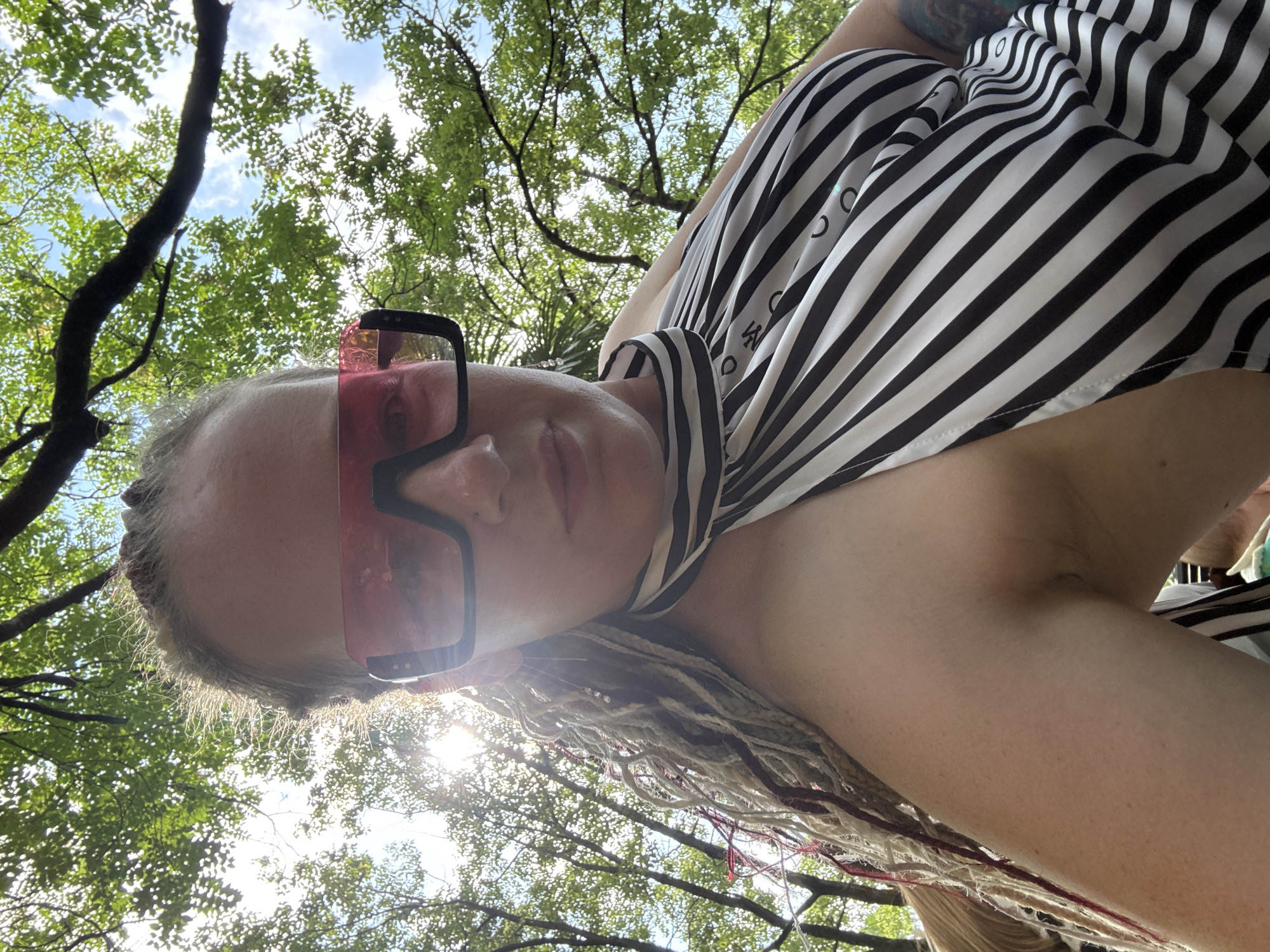 A woman with blonde dreadlocks and large pink sunglasses is pictured outdoors, looking up at the camera. She is wearing a black and white striped halter top. The background is dominated by green trees and a bright sky, suggesting a sunny day. The image appears to have been taken in Sochi, Russia, likely in a park or natural setting, given the foliage.