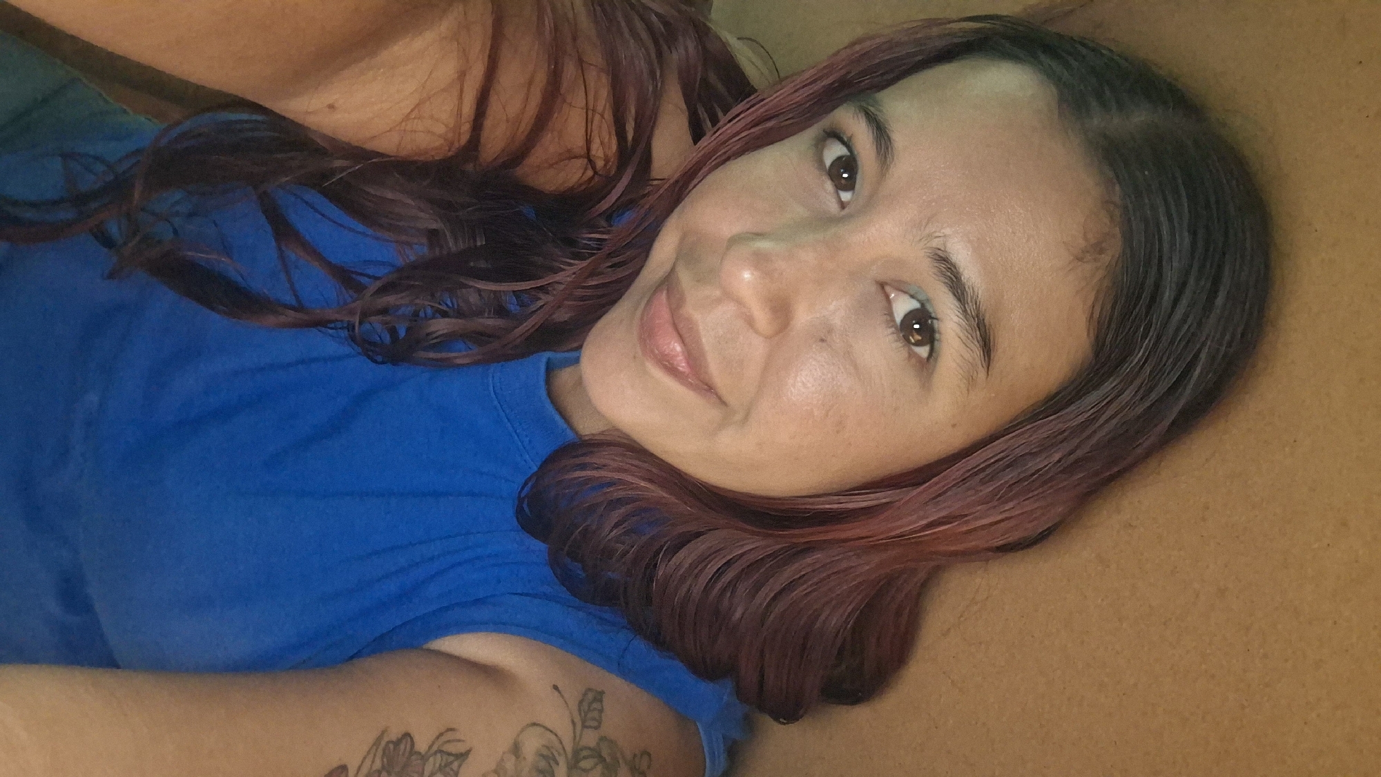 This is a close-up, overhead selfie of a woman lying down, with her head tilted. She has long, dark hair with reddish-pink streaks, and she is wearing a bright blue t-shirt. Her skin appears smooth and her eyes are looking directly at the camera with a gentle smile. There is a tattoo visible on her left arm. The background is a textured, brown surface. The lighting suggests it could be daytime, possibly indoors or in a shaded outdoor area, with a warm tone to the light. The overall mood is relaxed and intimate. The provided location context is Vallecito, Venezuela. There is no visible text in the image.