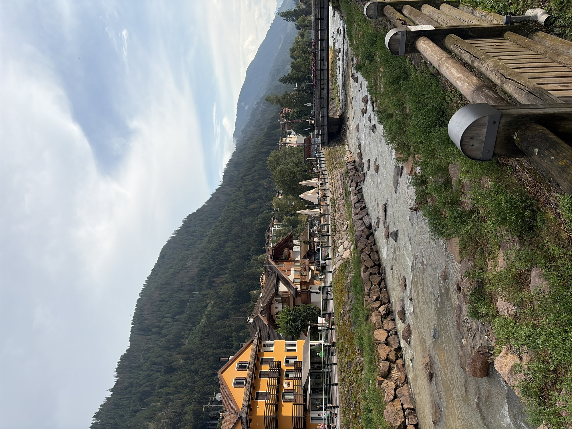 This is a medium-shot, eye-level, outdoor, horizontal photograph of the F Fontana stream in Moena, Italy, on a slightly overcast day. The stream, with its rocky bed and clear, flowing water, occupies the foreground and middle ground. On the left bank of the stream, a paved walkway runs alongside a row of buildings. The buildings, with their yellow and brown facades, are multi-story structures with balconies and pitched roofs. A wooden fence with metal caps separates the walkway from the stream. On the right bank, a similar wooden fence runs along the stream's edge, backed by green vegetation. In the background, a dense forest covers the steep slopes of a mountain range, and a bridge spans the stream in the distance. Several people are visible walking on the walkway and near the buildings. The sky is a pale blue with scattered clouds.