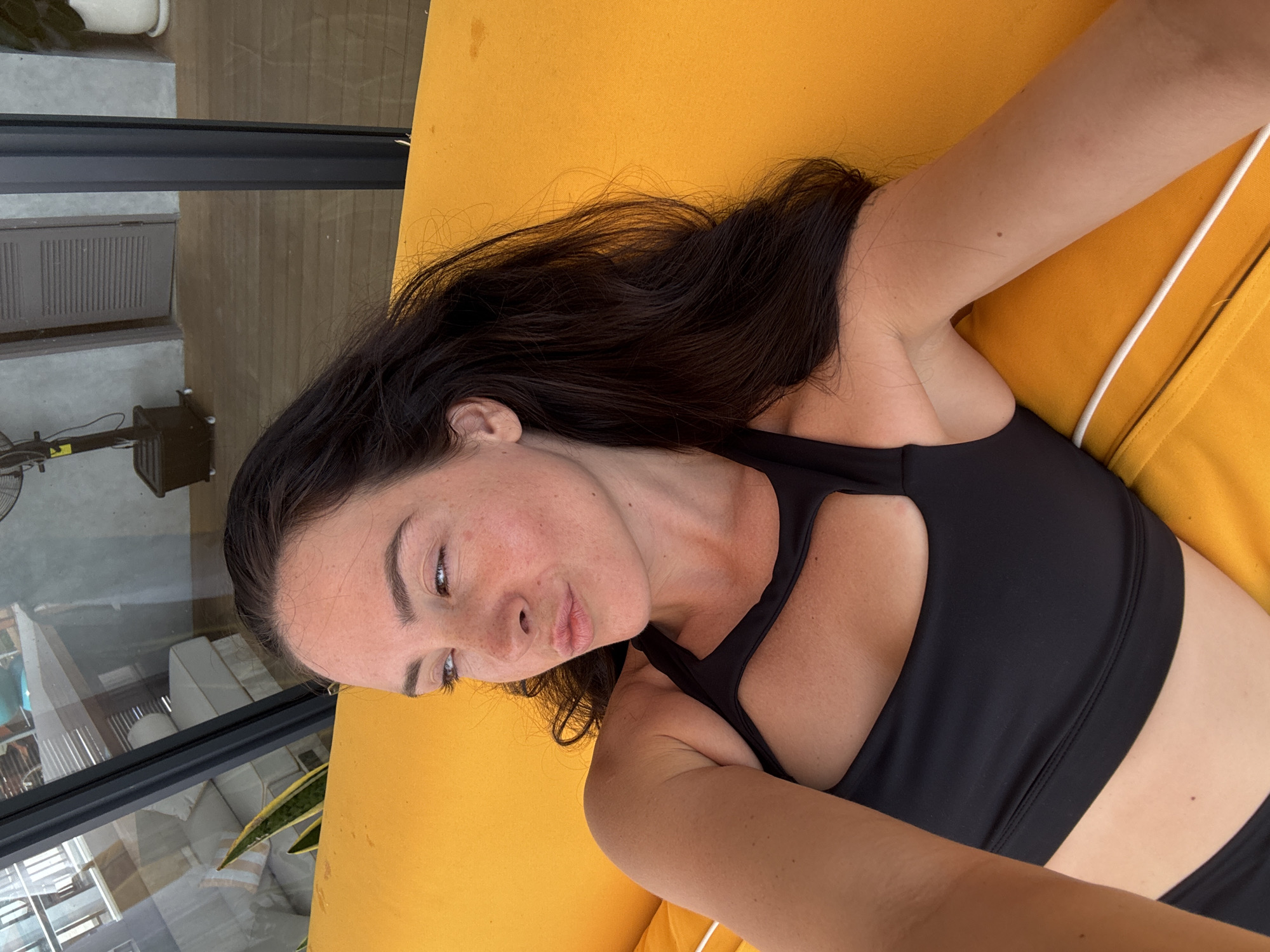 A woman with long, dark hair and fair skin with freckles lies on a bright yellow outdoor couch. She is wearing a black athletic top with a cutout detail at the chest. Her eyes are partially closed and looking to her left. Her lips are slightly pursed. The couch is outdoors, and in the background, through a glass-paneled wall, a lounge area with white furniture and a wooden floor is visible. A tall, dark fan on wheels is also present in the background. The scene appears to be in a resort or private residence in North Kuta, Indonesia, judging by the architecture and outdoor furnishings suggestive of a tropical climate.