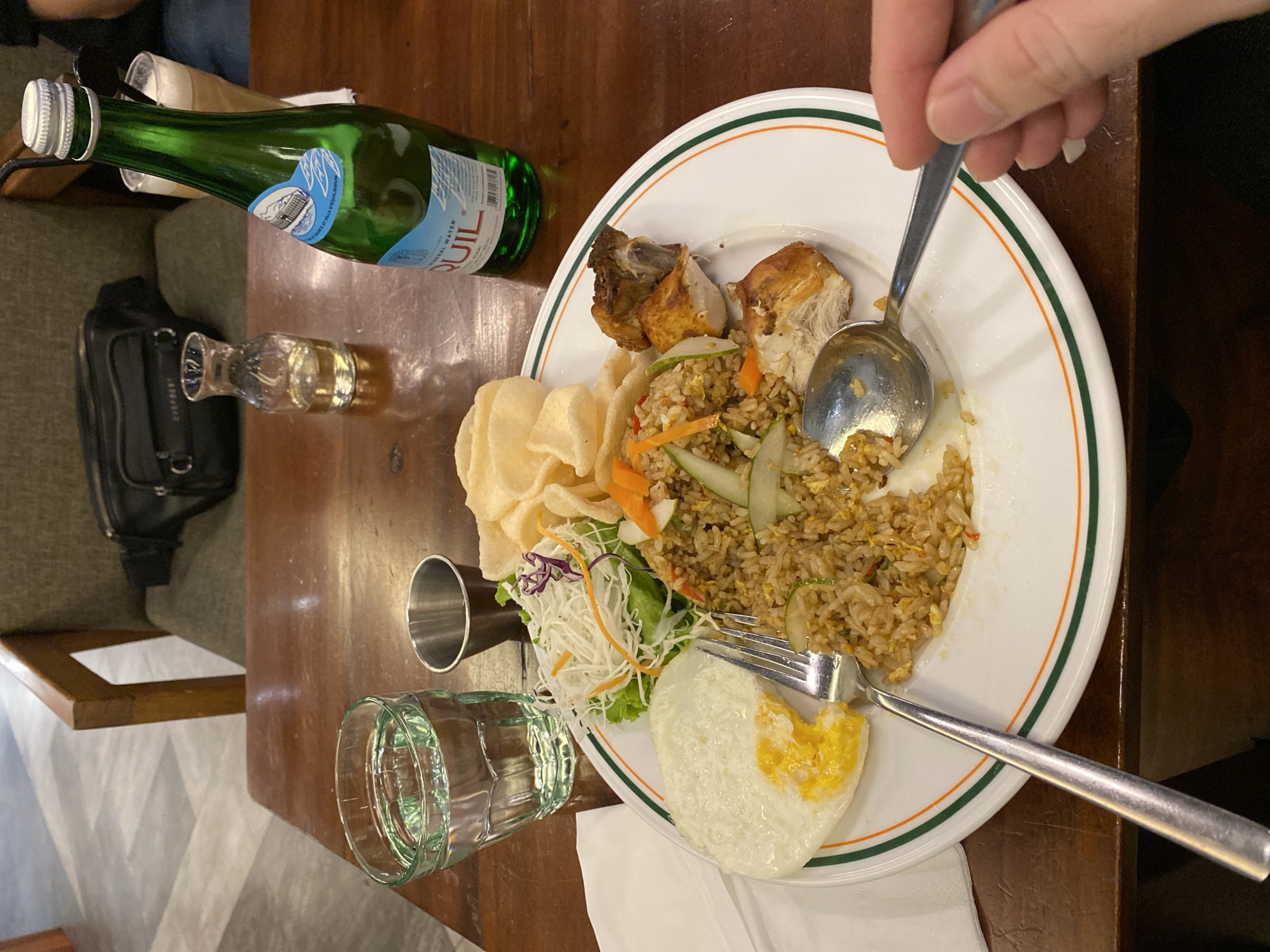 A close-up, eye-level shot captures a plate of Indonesian fried rice, chicken, and a fried egg on a wooden table. The fried rice is the centerpiece, surrounded by pieces of roasted chicken, a sunny-side-up egg with a visible yolk, a small salad, and crispy crackers. A fork rests on the plate, and a spoon is held by a person's hand, reaching towards the food. To the left, a glass of water and a small metal container are visible. A green bottle of mineral water stands to the right, partially obscuring a glass with a beverage. In the background, a chair with a dark bag slung over it and a small glass decanter suggest a dining setting. The overall impression is of a meal being enjoyed in a restaurant, possibly located in Banjarmasin, Indonesia, given the typical Indonesian cuisine depicted.