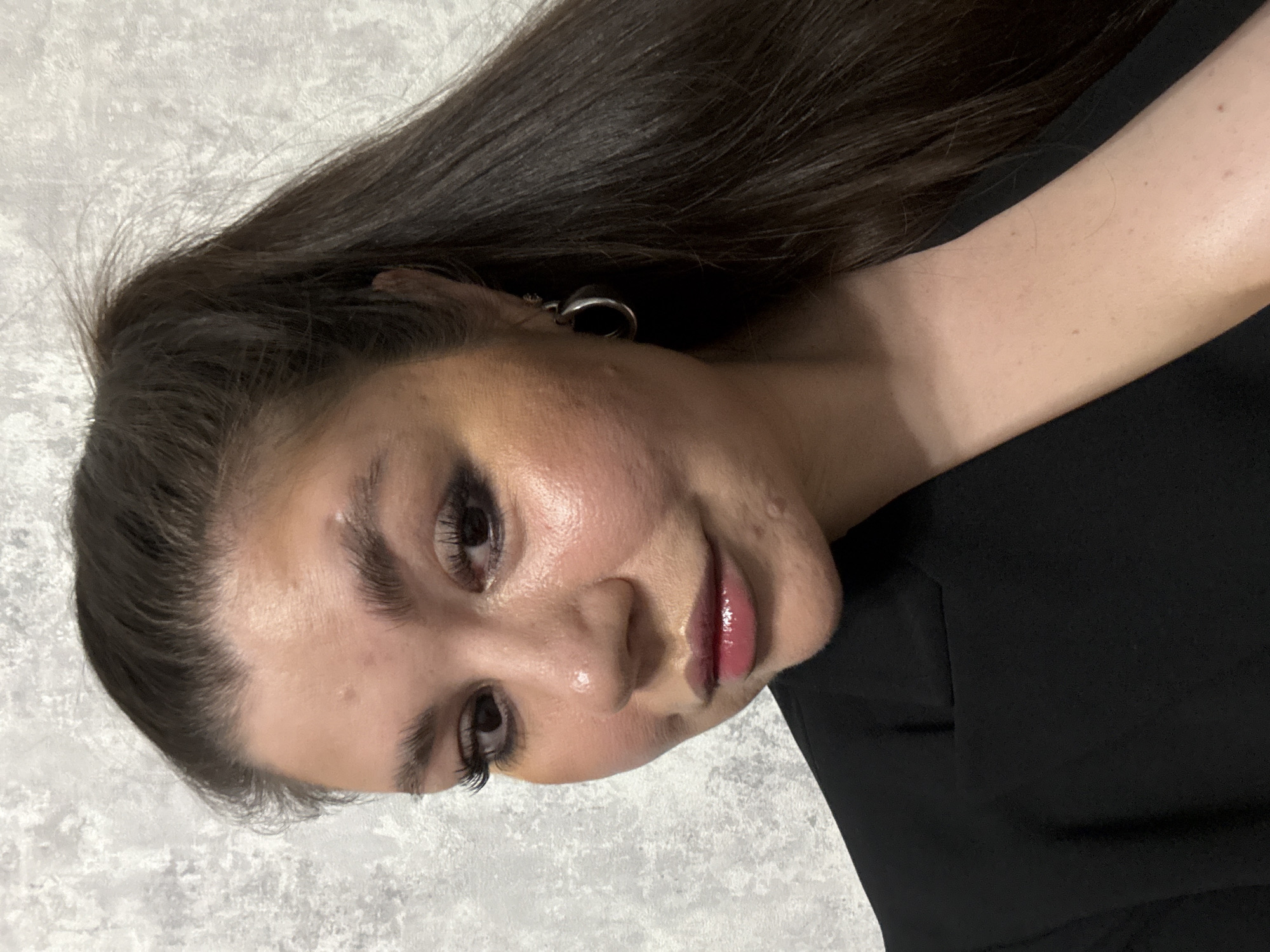 A close-up portrait of a young woman with long, dark brown hair. She has fair skin and is wearing dark eye makeup and glossy lip color. An earring is visible in her left ear. She is wearing a black garment. The background appears to be a textured, light gray surface. There is no information present in the image to indicate a location in Votkinsk, Russia.