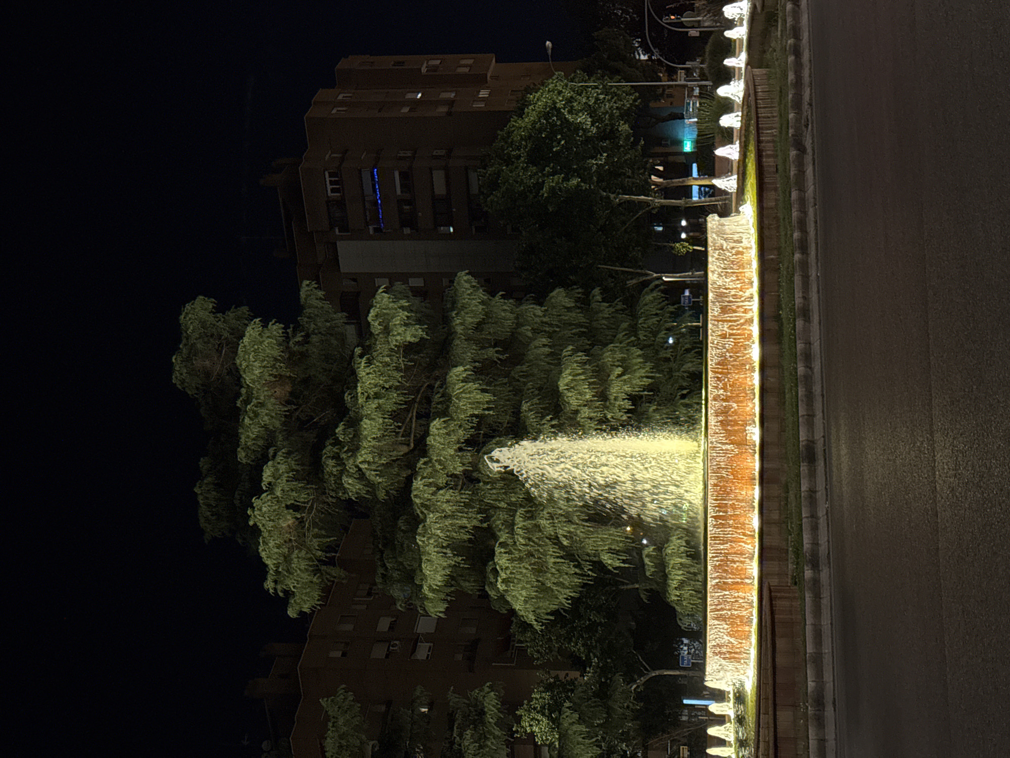 The image shows a brightly lit fountain in the center of a roundabout at night, with several trees and apartment buildings in the background. The fountain has a central plume of water illuminated by yellow lights, and a circular basin lined with orange lights, from which water cascades down. Smaller, illuminated water features are visible along the perimeter of the roundabout. The apartment buildings are dark, with some windows illuminated. The street in the foreground is wet, reflecting the lights. The scene is likely a public space in Madrid, Spain, given the context provided.