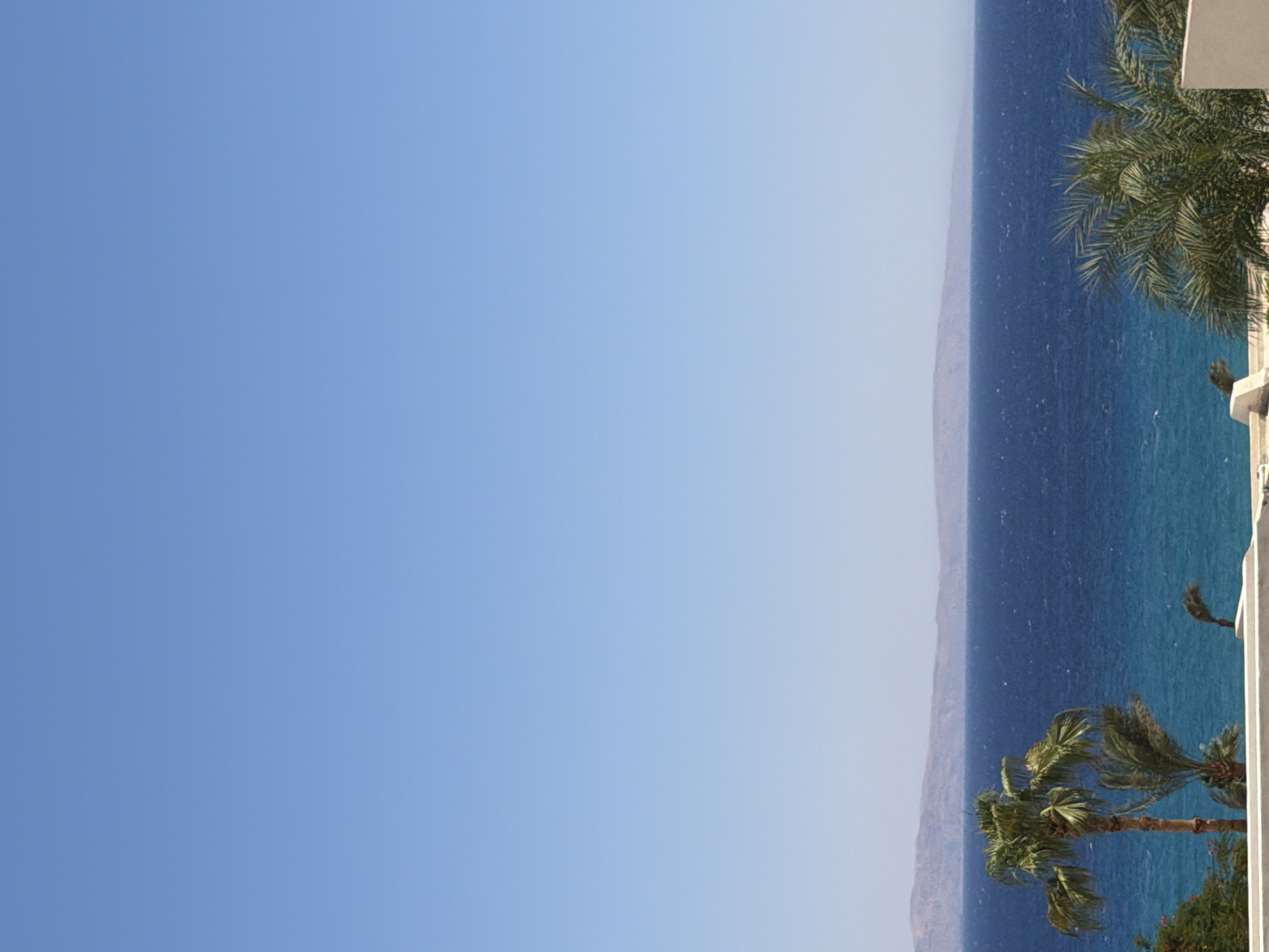 This is a landscape photograph taken from a slightly elevated vantage point, likely a balcony or rooftop, in Dahab, Egypt. The scene captures a clear, cloudless blue sky dominating the upper portion of the frame. Below the sky, a broad expanse of deep blue water, presumably the Red Sea, stretches across the middle ground. The water exhibits some texture, suggesting a gentle breeze or ripples on its surface.
In the distance, a hazy, low-lying landmass forms the horizon, its muted colors suggesting mountains or hills. In the foreground, a white, low-rise structure, likely part of a building or resort, is partially visible along the bottom edge of the image. To the left and center of the foreground, several palm trees are prominent. One mature palm tree stands tall with its fronds reaching upwards, while another smaller palm is situated behind it. To the far right, another palm tree is visible, its fronds appearing windblown. The presence of palm trees and the sea strongly suggests a coastal location in Dahab. The overall impression is one of a serene, sunny day in a tropical or subtropical coastal environment.