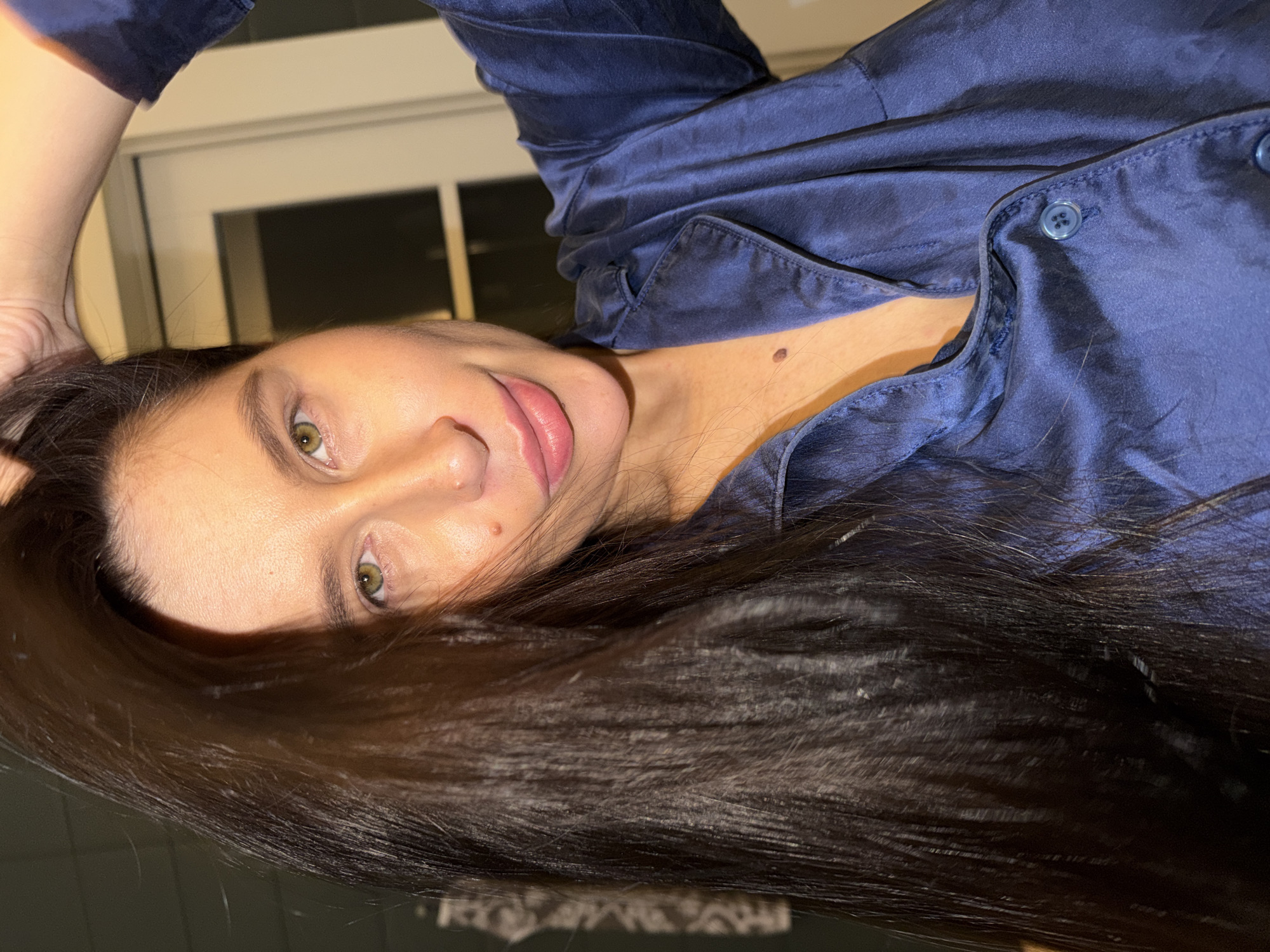 The image shows a person, likely female, reclining indoors in what appears to be a residential setting in Khimki, Russia. The individual is looking directly at the camera with a calm, relaxed expression and a subtle, soft smile.
The subject has long, dark brown hair that spreads across a significant portion of the image. They have green eyes, soft pink lips, and a light skin tone. Notable details on their skin include a small mole visible on the chin/jawline and another on the upper chest area, near the collarbone. One of their arms is raised, with fingers gently touching their hair above their head.
They are wearing a dark blue, satin or silk-like button-down shirt, which resembles a pajama top. The fabric has a sheen that catches the light, and blue buttons are visible down the front of the shirt.
The background is dimly lit with warm artificial light, suggesting the time of day is evening or night. A window with a horizontal divider is partially visible in the background. The overall scene conveys a sense of intimacy and tranquility. There is no visible text in the media file.