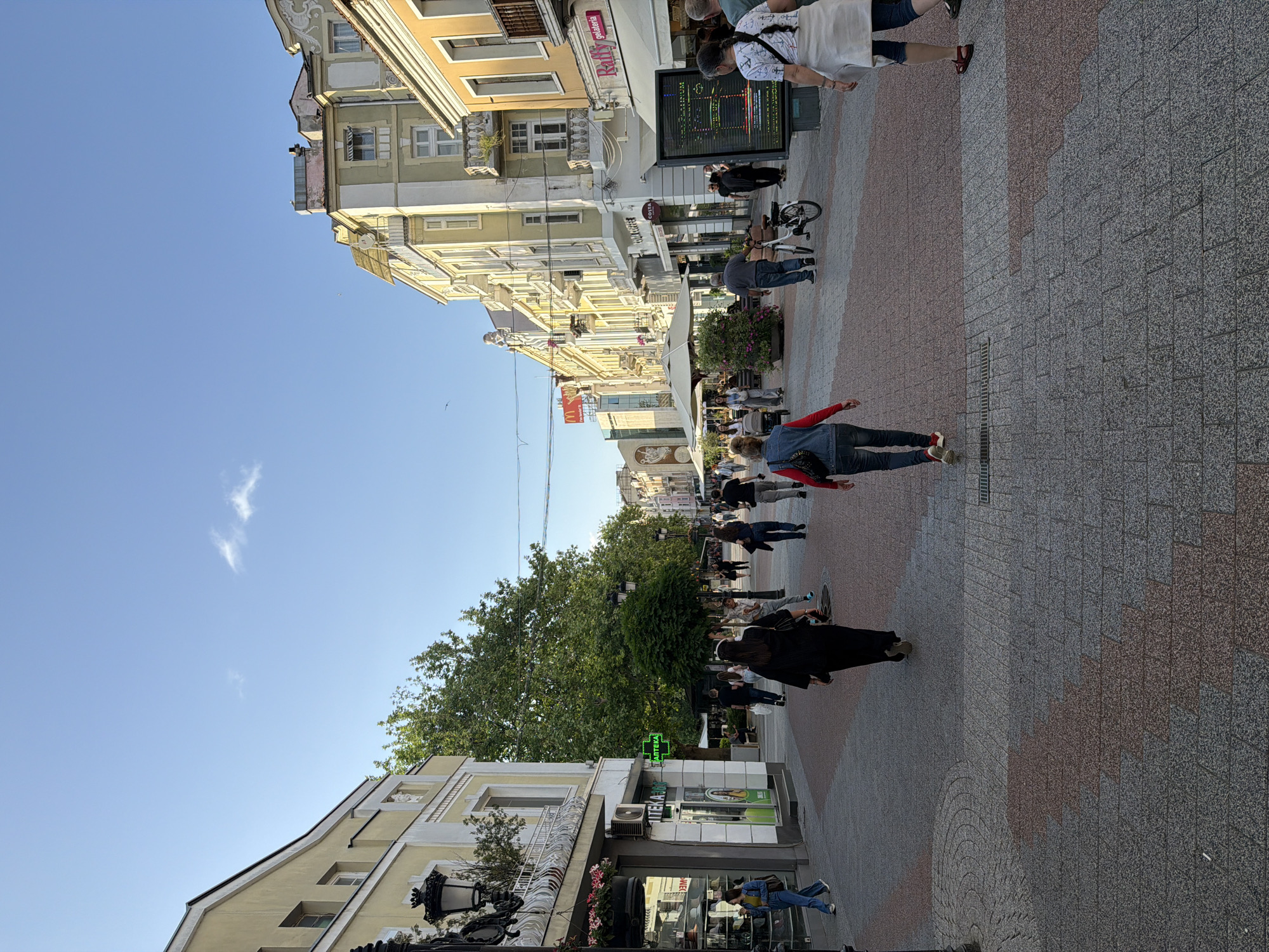 The image shows a city street in Unknown, Unknown. The street is paved with gray and red interlocking stones, and is lined with buildings of various heights and colors, mostly cream and yellow. A dense line of trees runs down the center of the street, providing shade for pedestrians. People are walking along the street, including a woman in a red jacket and blue jeans, a person in a black cloak, and a group of people near a large digital screen. There are several shops and businesses visible along the street, including a pharmacy with a green cross. The sky is clear and blue, with a few wispy clouds.