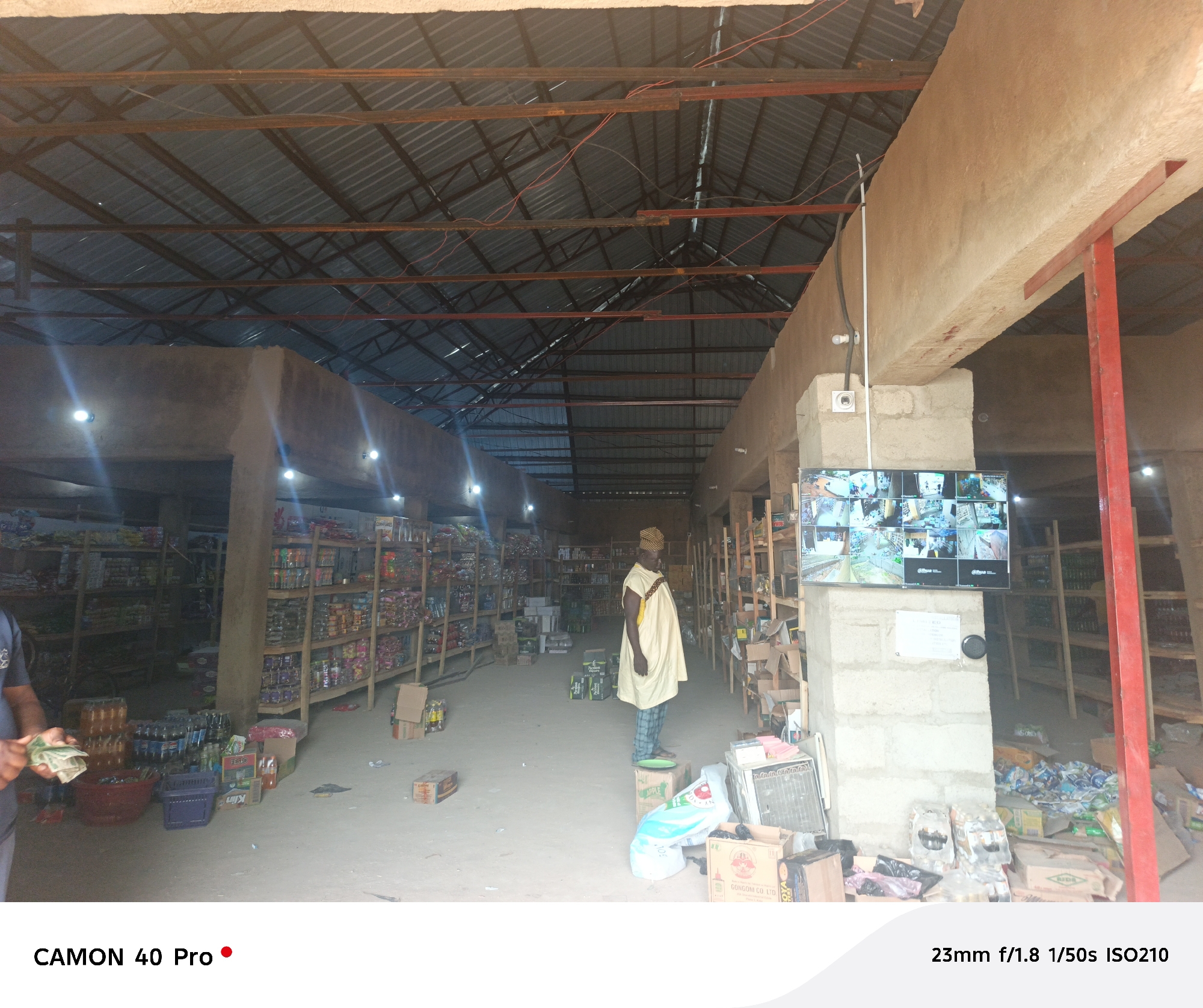 Here's a detailed description of the image:
**Setting:** The image is taken inside what appears to be a retail store or market stall, likely in Langtang, Nigeria. The building has a corrugated metal roof supported by a steel frame. Concrete pillars support a concrete beam running along the right side of the frame.
**People:**
* One man is clearly visible, wearing a cream-colored traditional outfit.
* Another person is partially visible on the left of the frame. They appear to be holding cash.
**Objects:**
* Shelves stocked with various goods line the sides of the building.
* Boxes and packages are scattered on the floor, particularly near the pillar on the right. Some visible brands include "FESTO" and "APPLE."
* A surveillance monitor is mounted on the concrete pillar to the right. The monitor displays feeds from multiple security cameras.
* There are two security cameras near the surveillance monitor.
* A red plastic basin and blue crate are located on the left.
**Actions/Interactions:**
* The man in the traditional outfit is standing, seemingly observing or perhaps waiting.
* The person on the left is presumably engaged in a transaction or handling money.
**Environment:**
* The lighting is a combination of natural light from outside and artificial lighting from bulbs inside.
* The store has a somewhat disorganized appearance.
* The location appears to be a marketplace, indicating a center of commerce and daily life.