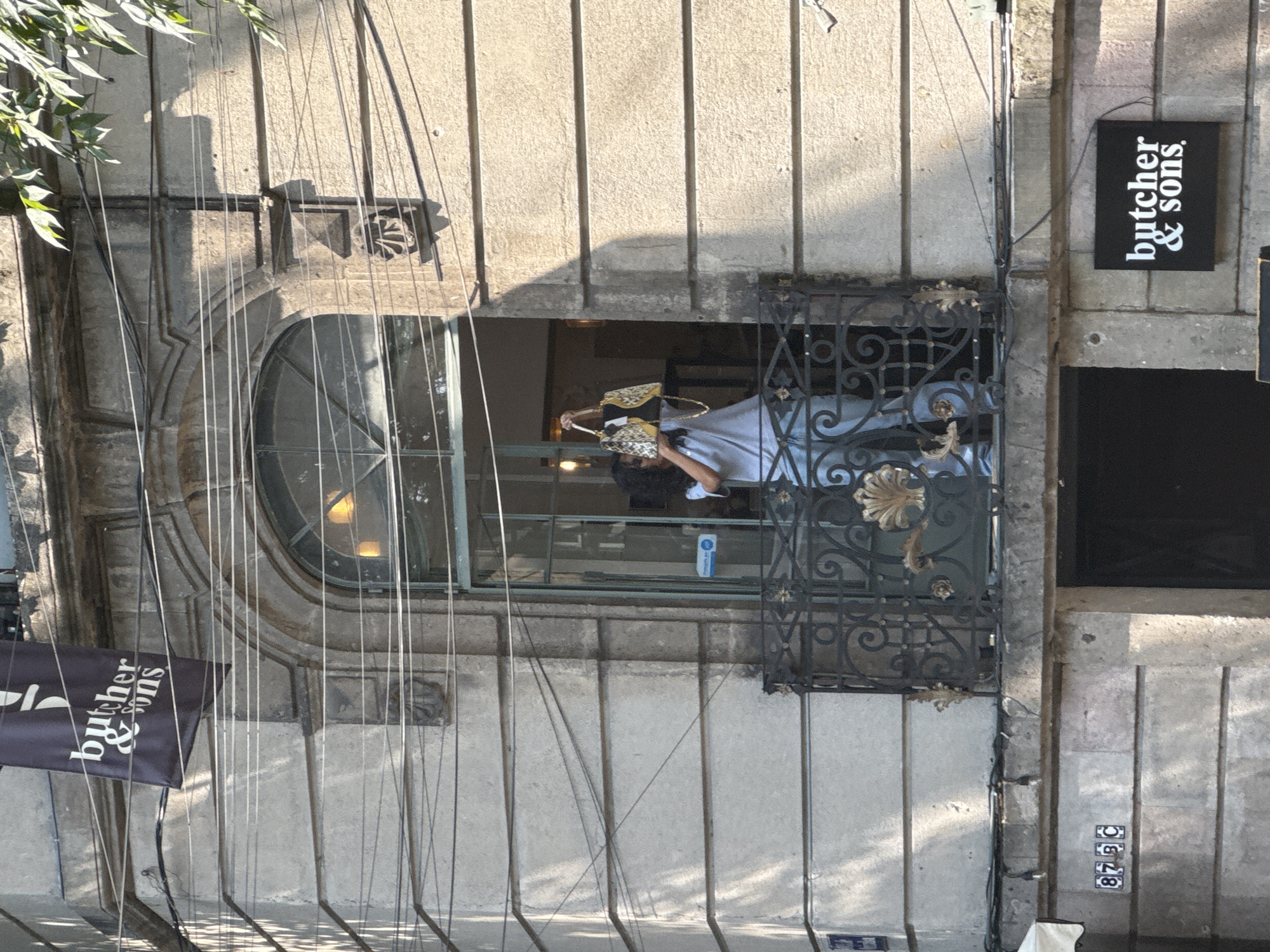 A medium shot photograph of the exterior of a building in Mexico City, Mexico, shows a person leaning out of an arched window. The window has an ornate metal railing. The person is holding a patterned bag. A sign for “butcher & sons” is visible on the right side of the building, and there is another sign for the same establishment hanging on the left. The building's address, “875C,” is visible on the right side. There are wires in the foreground.