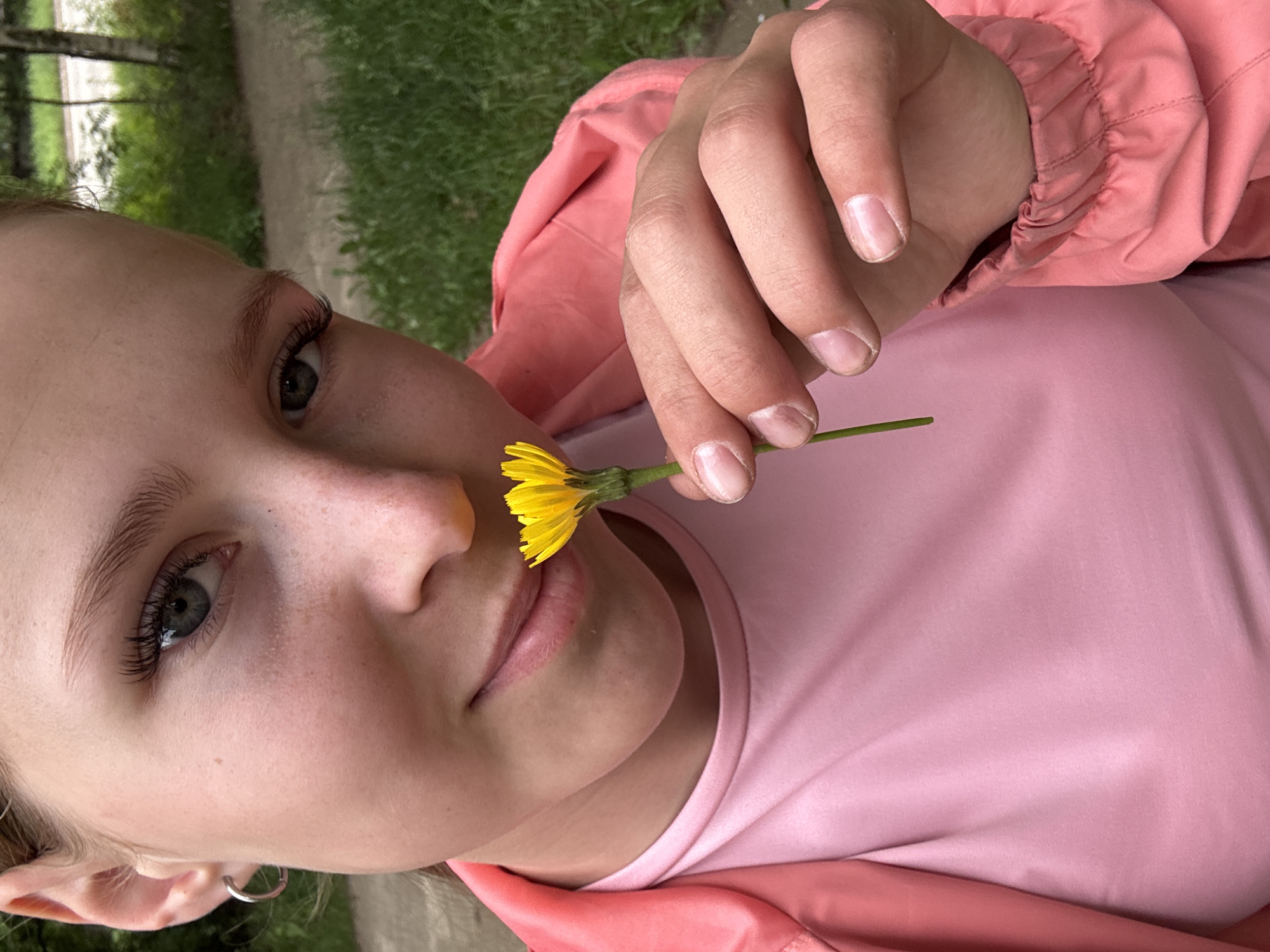 A young girl with blonde hair pulled back is shown in a close-up, outdoor portrait. She is wearing a pink t-shirt and a coral-pink jacket. Her left hand is holding a small yellow flower up to her nose, as if smelling it. The background is blurred, showing green grass and a pathway, suggesting a park or natural area within the city of Chaikovsky, Russia. The lighting is natural, indicating it is daytime.