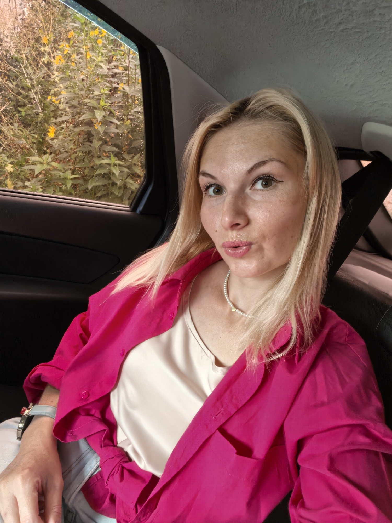 A blonde woman with light skin and light brown eyes is taking a selfie from the back seat of a car. She is wearing a bright pink button-up shirt over a cream-colored satin camisole, light-wash denim jeans, and a pearl necklace with a gold heart pendant. A silver watch with a light blue band is visible on her left wrist. Her lips are pursed. Through the car window to her left, a dense patch of green foliage with yellow flowers and water droplets is visible. The interior of the car, including the dark car door and a black seatbelt, is also visible. The scene is not identifiable as being in Sterlitamak, Russia.