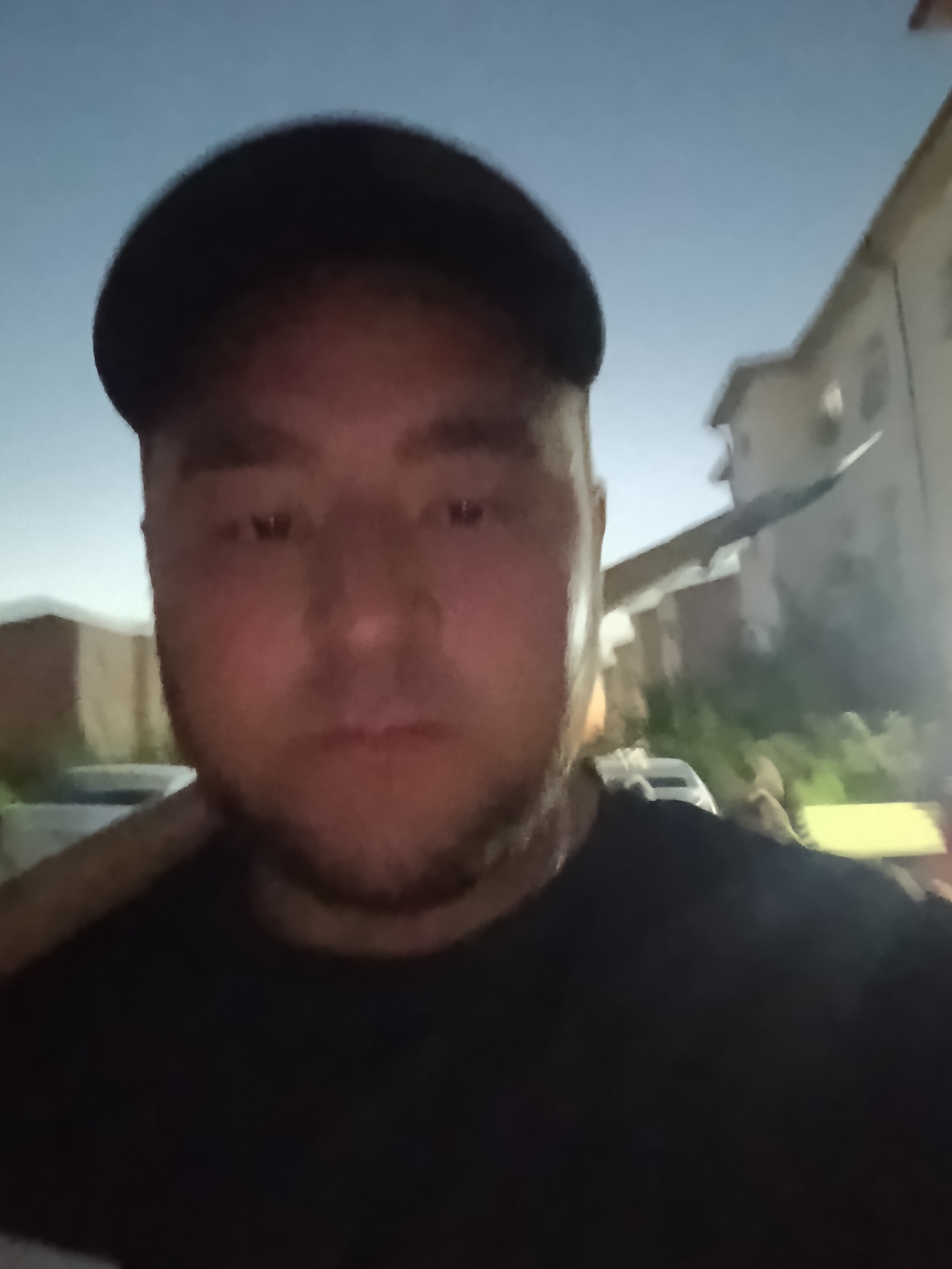 The image depicts a close-up selfie of a man wearing a black baseball cap and a black t-shirt. He has a beard and appears to be outdoors at dusk or night, with a dimly lit sky. In the background, there are out-of-focus buildings, some with lit windows, suggesting a residential or urban environment. A long, thin object, possibly a tool or a piece of equipment, extends diagonally across the upper right portion of the frame, originating from behind the man's head. The overall scene is grainy, indicating a low-light photograph. The location is identified as Zarafshan, Uzbekistan.