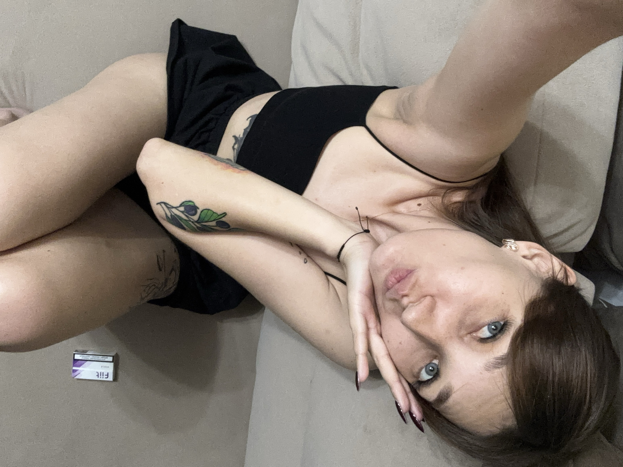 A young woman is lying on her back on a beige couch. She has blue eyes, dark brown hair, and light skin. She is wearing a black top and black bottoms. She has tattoos on her left arm and leg, and a small mole on her collarbone. She is holding her right hand up to her face, partially covering her mouth. A small package of "Fiit" cigarettes is on the couch to her left. The scene appears to be indoors, possibly in a home. The image does not contain information that would allow for determining the specific location within the city of Novokuznetsk, Russia.