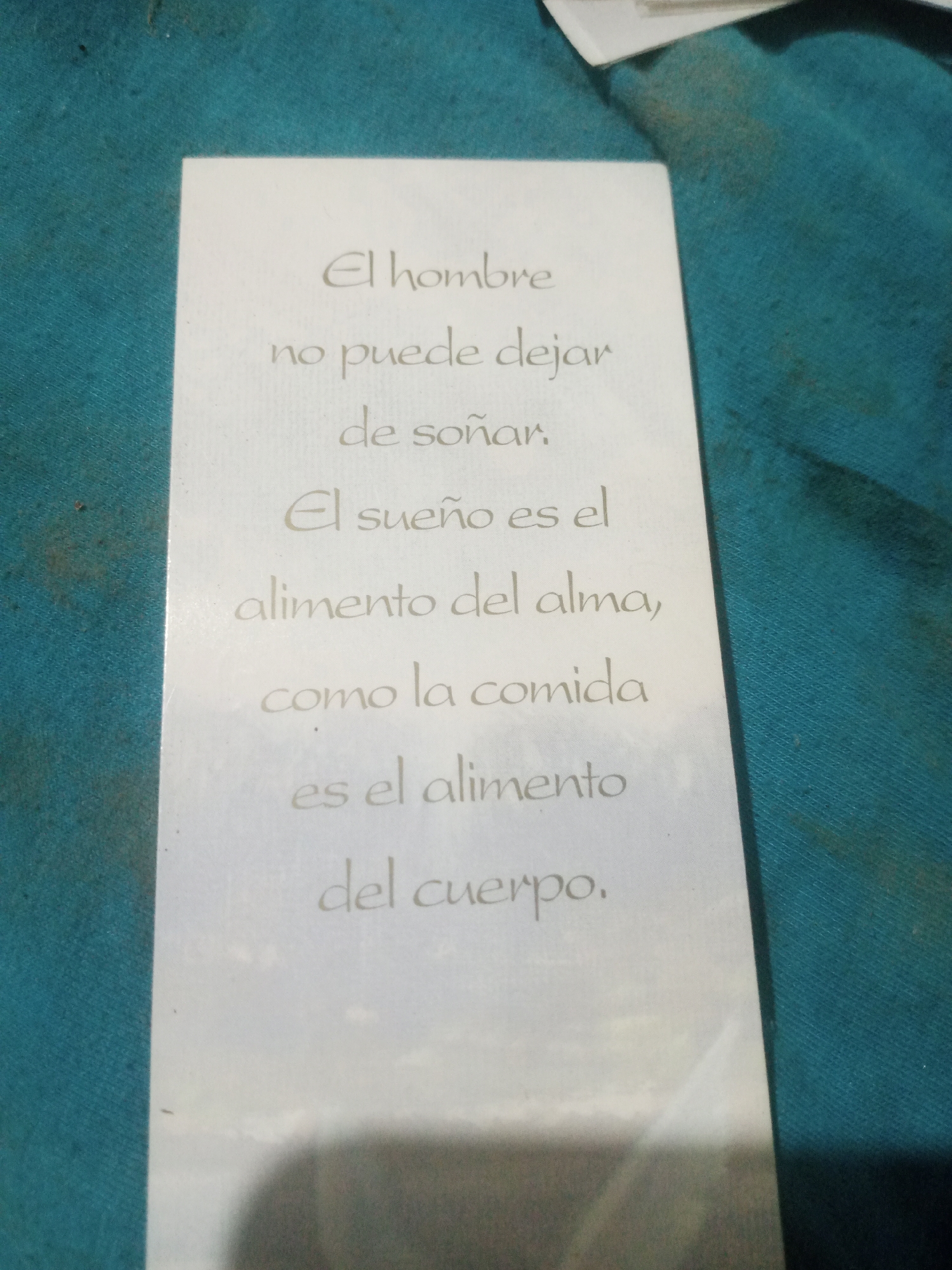 The image shows a white card with text written in a script font. The text appears to be a quote. The card is placed on a teal-colored textured surface, possibly fabric, with some dark smudges. The lighting in the image creates a shadow on the lower right portion of the card. The scene does not provide any information about the city of Santa Ana, Venezuela. The quote reads: "El hombre no puede dejar de soñar. El sueño es el alimento del alma, como la comida es el alimento del cuerpo."