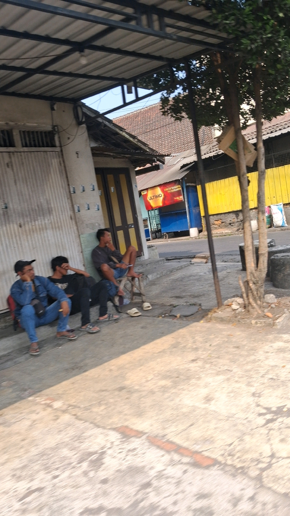 The image shows three men sitting outside a building with a covered porch. The building appears to be a shop or a small business, with a yellow and blue facade visible in the background. The men are casually dressed, and two of them are smoking. One man is sitting on a chair, while the other two are sitting on the ground. A tree is visible on the right side of the image, and the ground is paved with concrete. The scene appears to be a street in a town or city, possibly in Indonesia, given the context of Muntilan. The lighting suggests it is daytime.