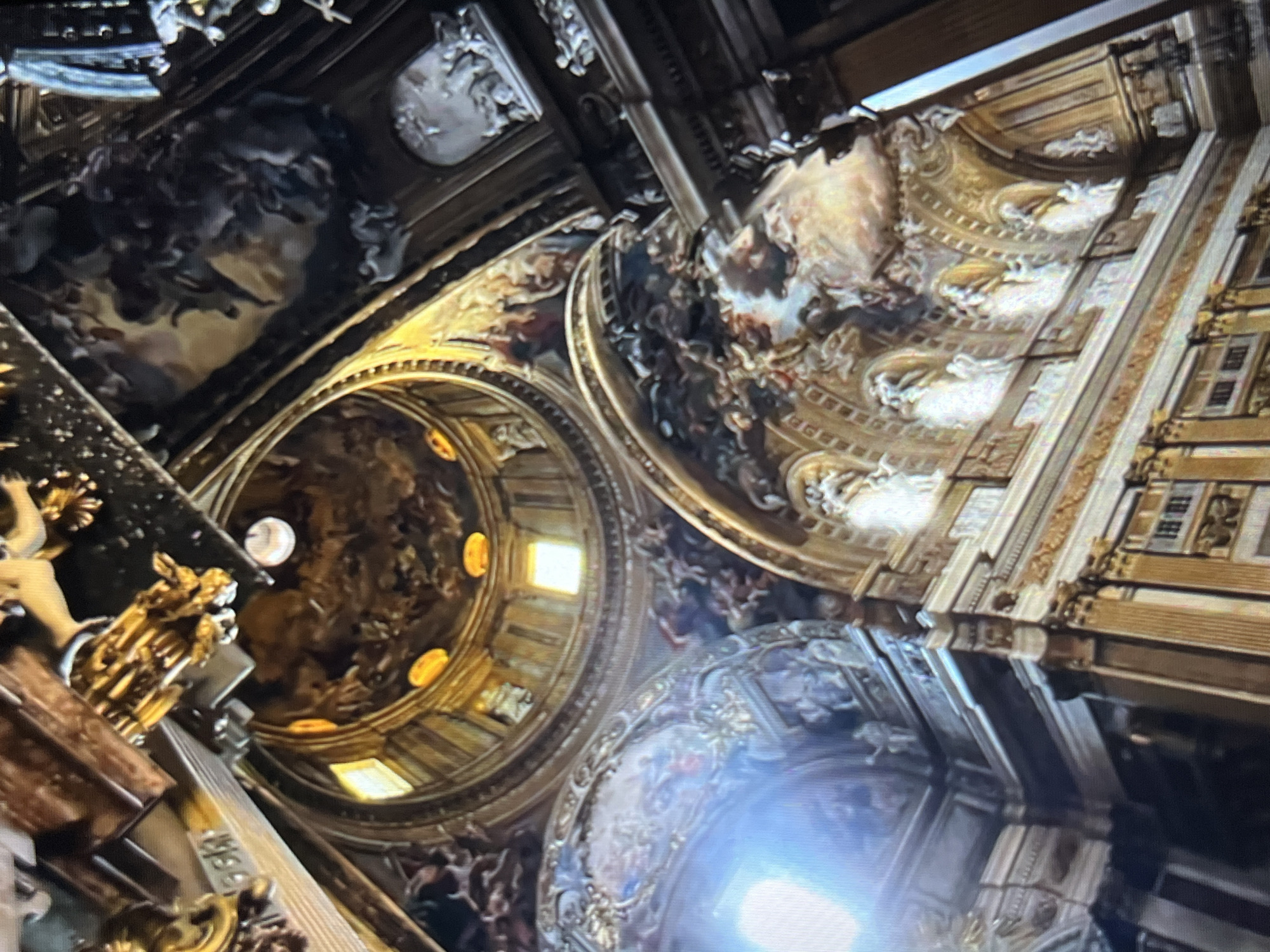 The image shows an upward-angled view of the elaborately decorated interior ceiling of a church. The ceilings are composed of domes and panels adorned with paintings, likely depicting religious scenes or figures. The color palette is dominated by golds, browns, and dark blues. The building's interior features architectural elements like arches, columns, and recessed panels. There are no indications of people in the image. The location is uncertain and, therefore, cannot be identified.