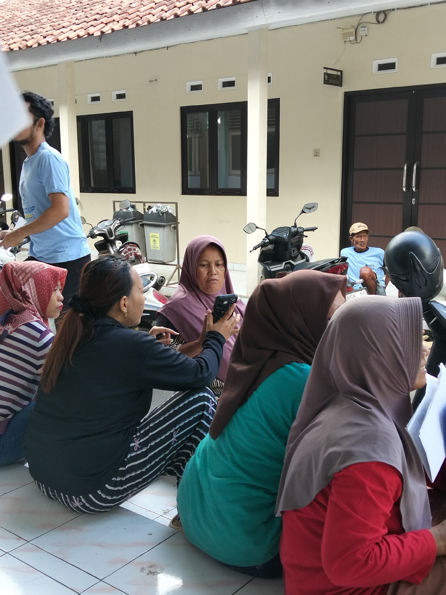 In an outdoor setting in Purwokerto, Indonesia, several individuals are gathered near a building with a tiled roof and cream-colored walls. A man wearing a blue t-shirt stands to the left, partially obscured, possibly in front of a motorcycle. In the foreground, four women are seated on the tiled ground, facing various directions. One woman in the center, wearing a purple headscarf, is looking at a smartphone she holds in her hand. Another woman to her left, wearing a red and white striped headscarf, is also looking towards the center. To the right, two women are seated, both wearing headscarves, with one in a teal-colored shirt and the other in a red shirt. Their interactions are not clearly defined beyond their proximity. Motorcycles and trash bins are visible in the background, suggesting a public or communal area. An older man in a light blue t-shirt and a tan cap is seated on a motorcycle further back, looking towards the left. The overall scene appears casual and relaxed, with people congregating outdoors.