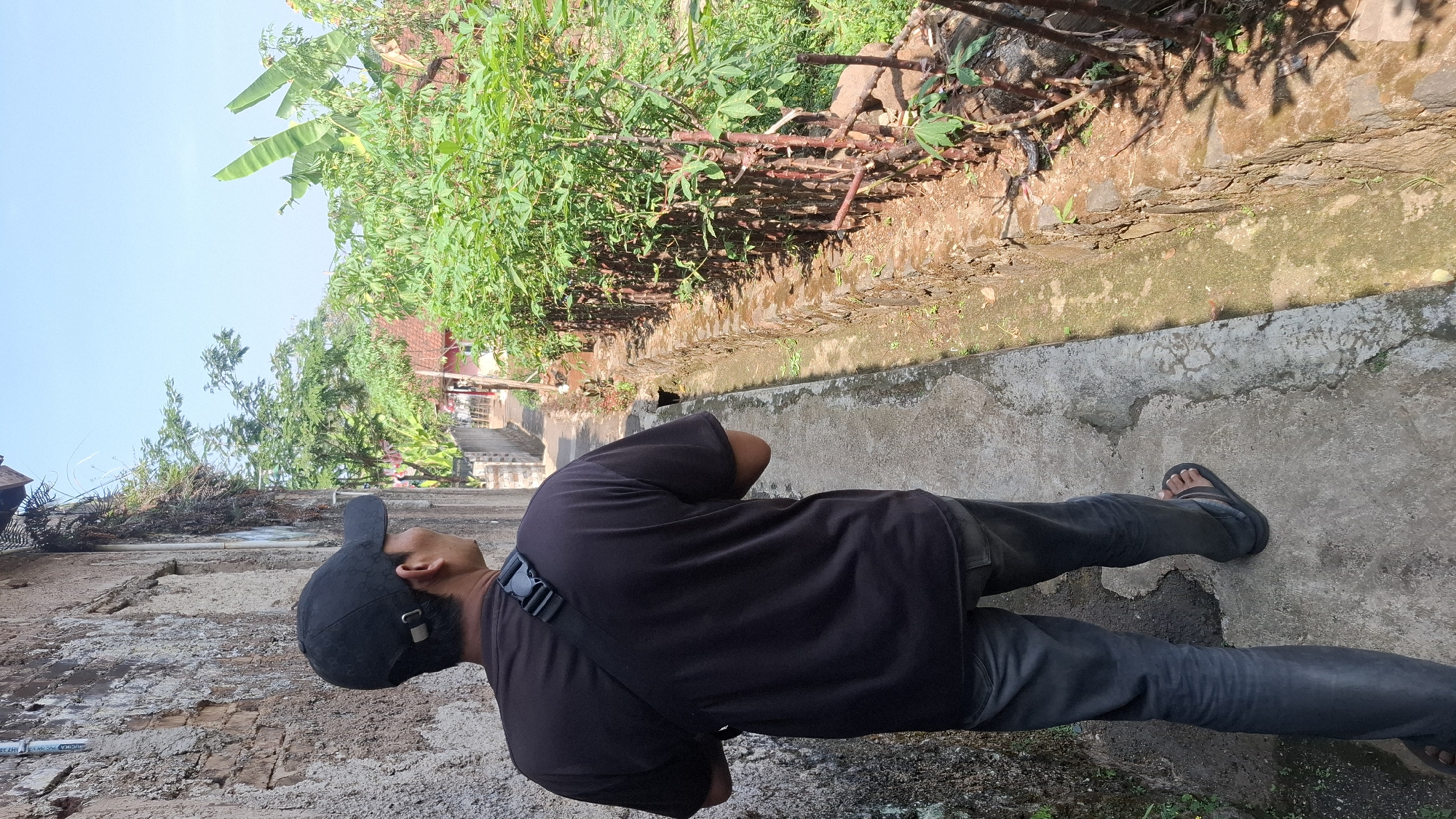 A person wearing a black t-shirt, black cap, and dark jeans walks down a narrow concrete pathway next to a weathered brick wall. The pathway is bordered on the right by a stone-lined drainage ditch with greenery growing along its edge, including several young trees with reddish stems. Beyond the ditch and greenery, a village scene unfolds with houses and banana trees visible under a clear blue sky. The overall setting appears to be a residential area in Cilaku, Indonesia, likely a rural or semi-urban environment, characterized by its weathered infrastructure and lush vegetation.