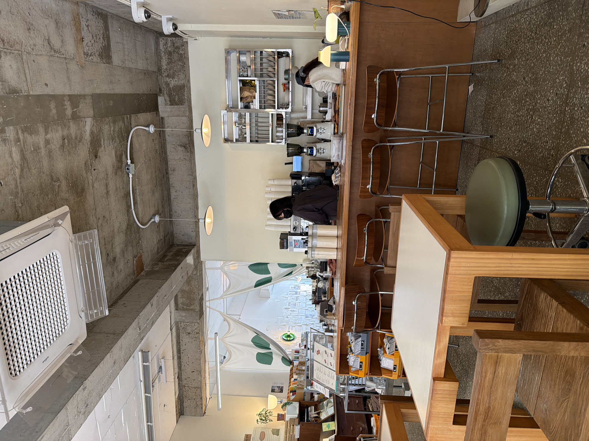 The image depicts the interior of a cafe or coffee shop, likely in Seoul, South Korea, judging by the architectural style and general ambience. The scene is captured from a high angle, providing a comprehensive view.
The cafe features a long wooden counter, behind which two individuals are working. One person, wearing a light-colored top, appears to be engaged in preparing beverages or food items. The other person, dressed in a darker outfit and wearing a face mask, is positioned behind the counter, possibly handling transactions or managing supplies. Coffee-making equipment, including espresso machines and grinders, are arranged on the counter. A set of metal shelves filled with cups, plates, and other utensils is located on the wall behind them.
Several high chairs with wooden seats and metal frames are placed along the counter for customers. A light-colored wooden table and a green stool are visible in the foreground.
The interior design incorporates a mix of materials, including exposed concrete on the ceiling and walls, which is juxtaposed with the warm tones of the wood used for the counter, tables, and chairs. Windows, partially covered with decorative white and green panels, provide natural light.
The space appears to be clean and orderly, with an emphasis on functionality and aesthetics. The overall atmosphere seems inviting and relaxing.