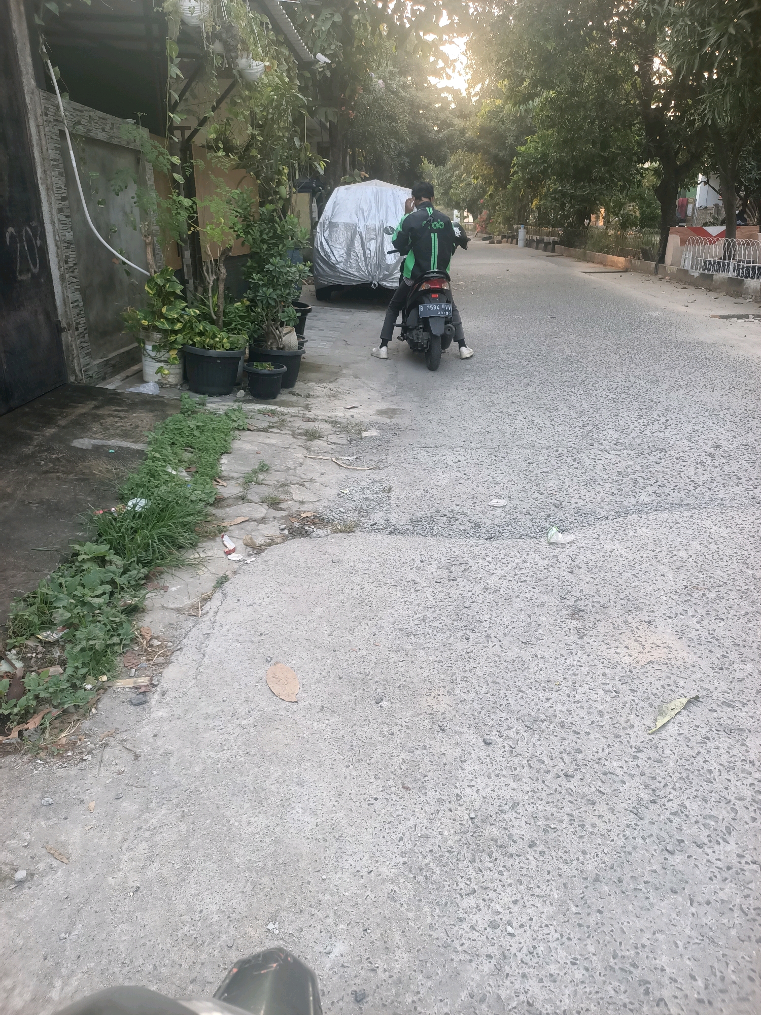 In Wanasari, Indonesia, a person wearing a green and black Grab uniform is riding a dark-colored motorcycle. The motorcycle is parked on a paved road, with a silver-colored car cover visible behind it. The area appears to be a residential street, with houses and trees lining the road. There are also several potted plants on the left side of the image. The sun is setting, casting a warm glow over the scene. There is some litter on the ground, including a plastic bottle. The motorcycle's license plate is visible and reads "B 2584 FVV".