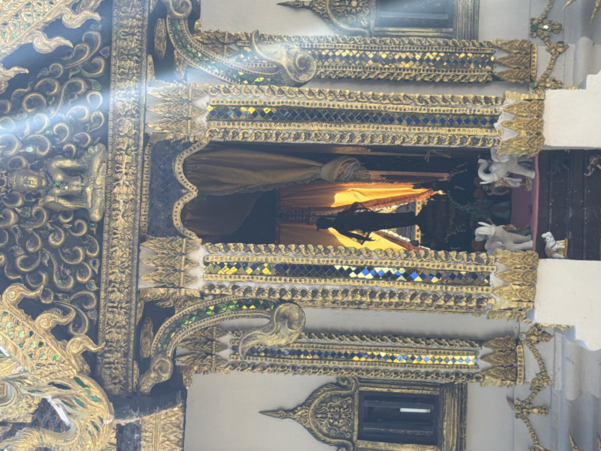 This image captures a close-up view of an ornate, likely religious structure, possibly a shrine or temple facade. The architecture is heavily decorated with intricate gold detailing, featuring scrollwork, floral motifs, and small figurines. Panels of blue and green mirrored tiles are interspersed within the golden ornamentation, adding a mosaic-like quality.
The central focus is an open archway revealing a darker interior, draped with golden-yellow fabric. A silhouetted figure, appearing to be a statue or effigy of a standing person, is visible within this space, illuminated by a bright light source from behind, creating a dramatic silhouette. Below the archway, several small white elephant figurines are placed, some standing and some with their trunks raised.
Sunlight streams into the scene from the upper right, casting bright beams across the gilded surfaces and highlighting the textures and details of the architecture. The overall impression is one of opulence and spiritual reverence. The materials appear to be wood, metal (likely gilded), and possibly stone or plaster for the base structure. The environment suggests an outdoor setting, given the natural sunlight.