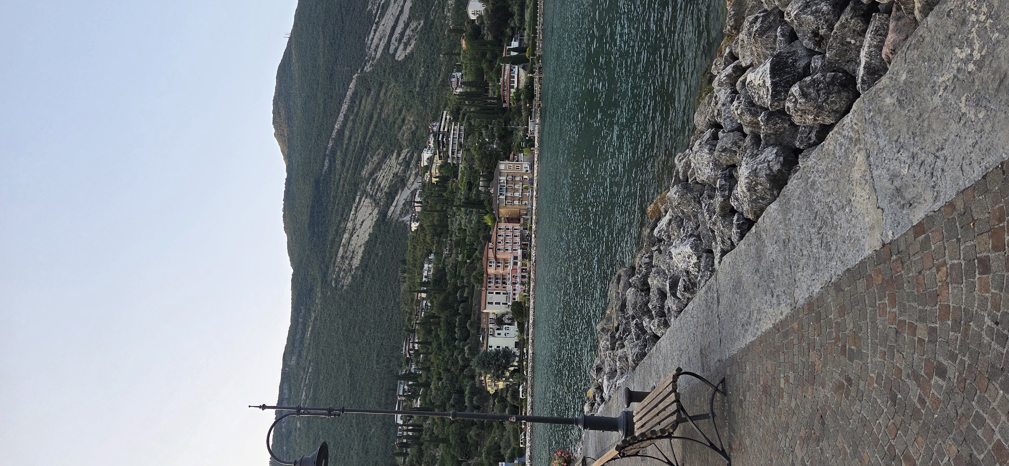 The image depicts a scenic view of the lakefront in Torbole, Italy. In the foreground, a stone embankment lines the water, with a paved walkway made of reddish-brown cobblestones. A weathered wooden bench with metal legs is placed on the walkway, next to a black lamppost. The lake, a deep blue-green, extends towards the background, where a lush green mountainside rises. Dotted along the hillside are various buildings, some appearing to be hotels or residences, with colorful facades. The sky above is a clear, pale blue, with a hint of haze. The overall impression is one of tranquility and natural beauty.