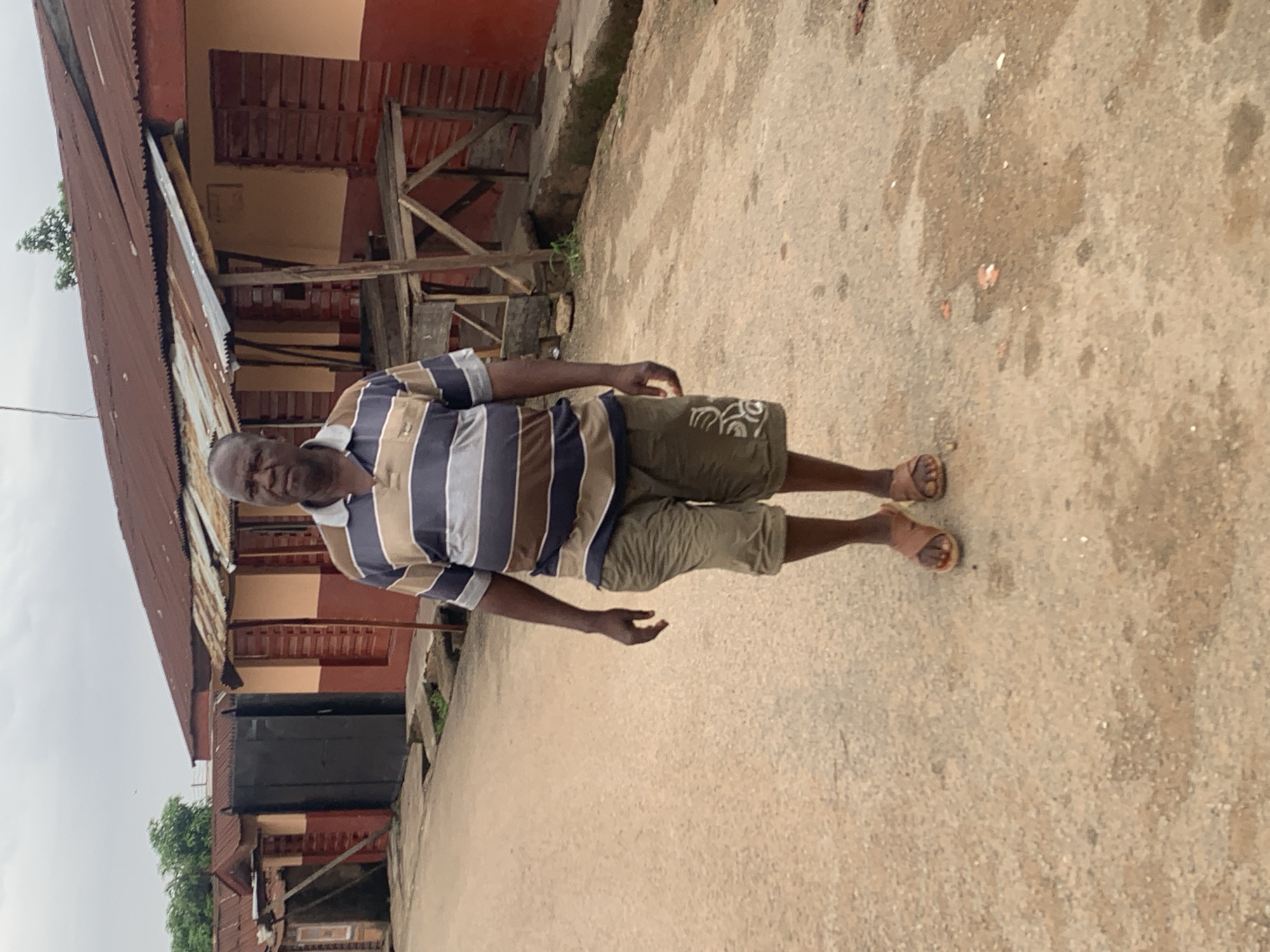 The photograph shows an older man standing on a paved surface next to a building with a rusted corrugated metal roof. The building has two stories with red and orange paint. The man is wearing a striped shirt, olive green shorts, and sandals. The location is identified as Jos, Nigeria.