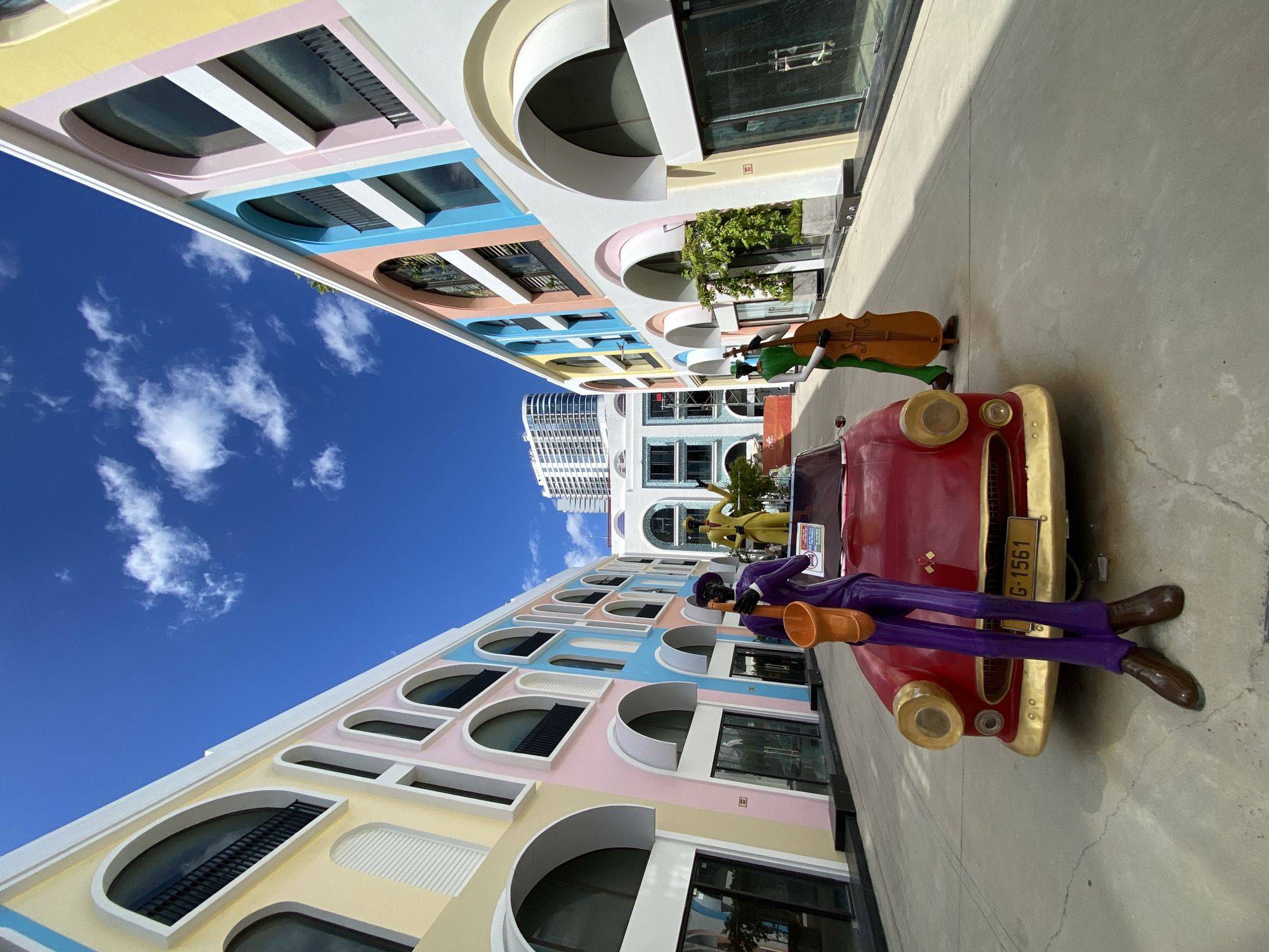 The image captures a vibrant street scene in Nha Trang, Vietnam, with a focus on the colorful architecture and artistic installations. The buildings on either side of the street are painted in a pastel palette of yellow, pink, blue, and white, featuring arched windows and balconies. In the foreground, a bright red vintage car serves as a centerpiece. Two human-like statues, dressed in purple and green, are positioned near the car; the one in purple holds a saxophone, while the one in green stands with a cello. A larger, yellow-clad statue is visible further down the street. The sky above is a clear blue with scattered white clouds. The street appears to be a pedestrian area or a plaza designed for artistic display and public enjoyment, likely part of a resort or entertainment complex in Nha Trang.