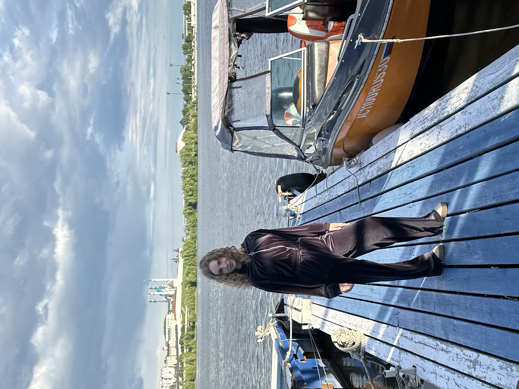 A young woman in a dark velvet tracksuit is lying on a blue wooden dock. She has blonde curly hair and is facing the camera. Behind her is a river and a cityscape, which includes the Qolşärif Mosque in Kazan, Russia. A boat is docked next to the woman, partially obscuring the view of the river. The sky is cloudy.