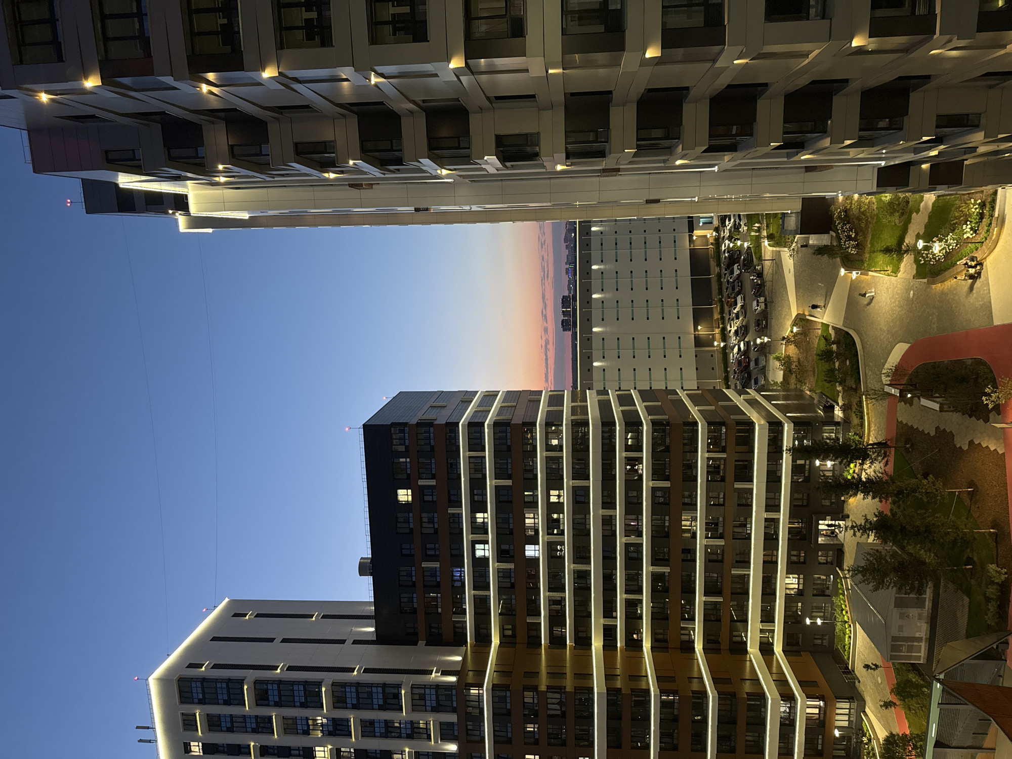 This is a medium shot, outdoor, eye-level, vertical photograph of a modern residential complex at dusk in Novosibirsk, Russia. The sky is a deep blue, transitioning to a soft pink and orange hue near the horizon. The scene is dominated by two tall, contemporary buildings with illuminated facades, their windows reflecting the fading light. The building on the left is taller and appears to be clad in white and dark brown material, with horizontal bands of bright white lights outlining its structure. The building on the right is also tall, with a lighter colored facade and vertical accent lighting. Between these buildings, a courtyard area is visible, featuring paved walkways, green spaces, trees, and outdoor lighting. A parking lot filled with cars is visible in the background, as is a long, low-rise industrial or commercial building. Several indistinct figures are present in the courtyard area, walking or sitting. The overall impression is of a well-lit, modern urban environment.