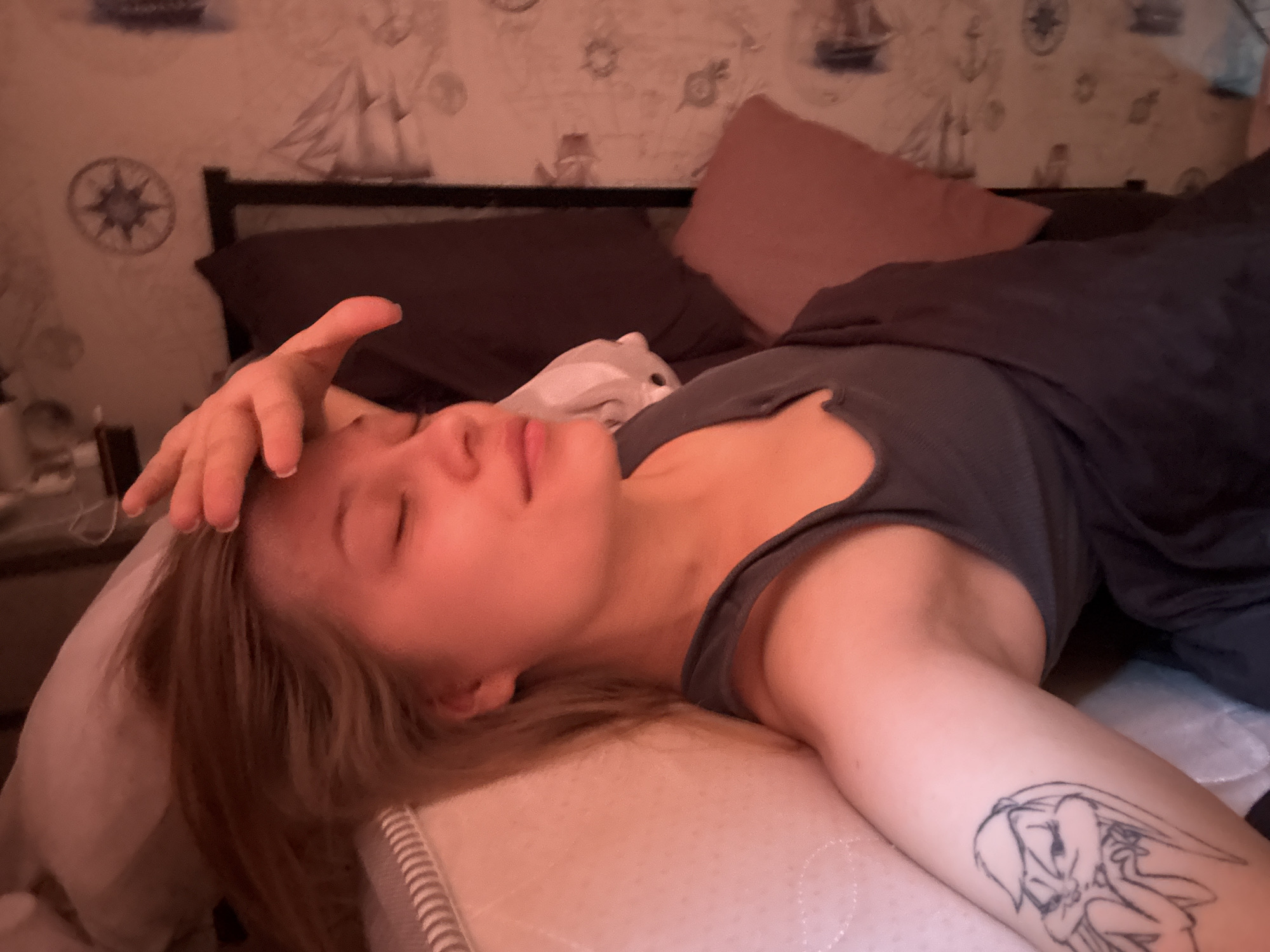 A woman with light skin and light brown hair is lying on a bed. She is wearing a gray tank top and is partially covered by a dark gray blanket. Her eyes are closed, and her right hand is resting on her forehead. On her left arm is a tattoo of Bugs Bunny. The bed has a dark gray headboard, and two pillows are visible: a dark gray pillow and a pink pillow. The wallpaper behind the bed has a nautical theme, with drawings of sailboats, compasses, and anchors. The scene appears to be indoors, possibly in a bedroom. The location is the city of Chaikovsky, Russia.
