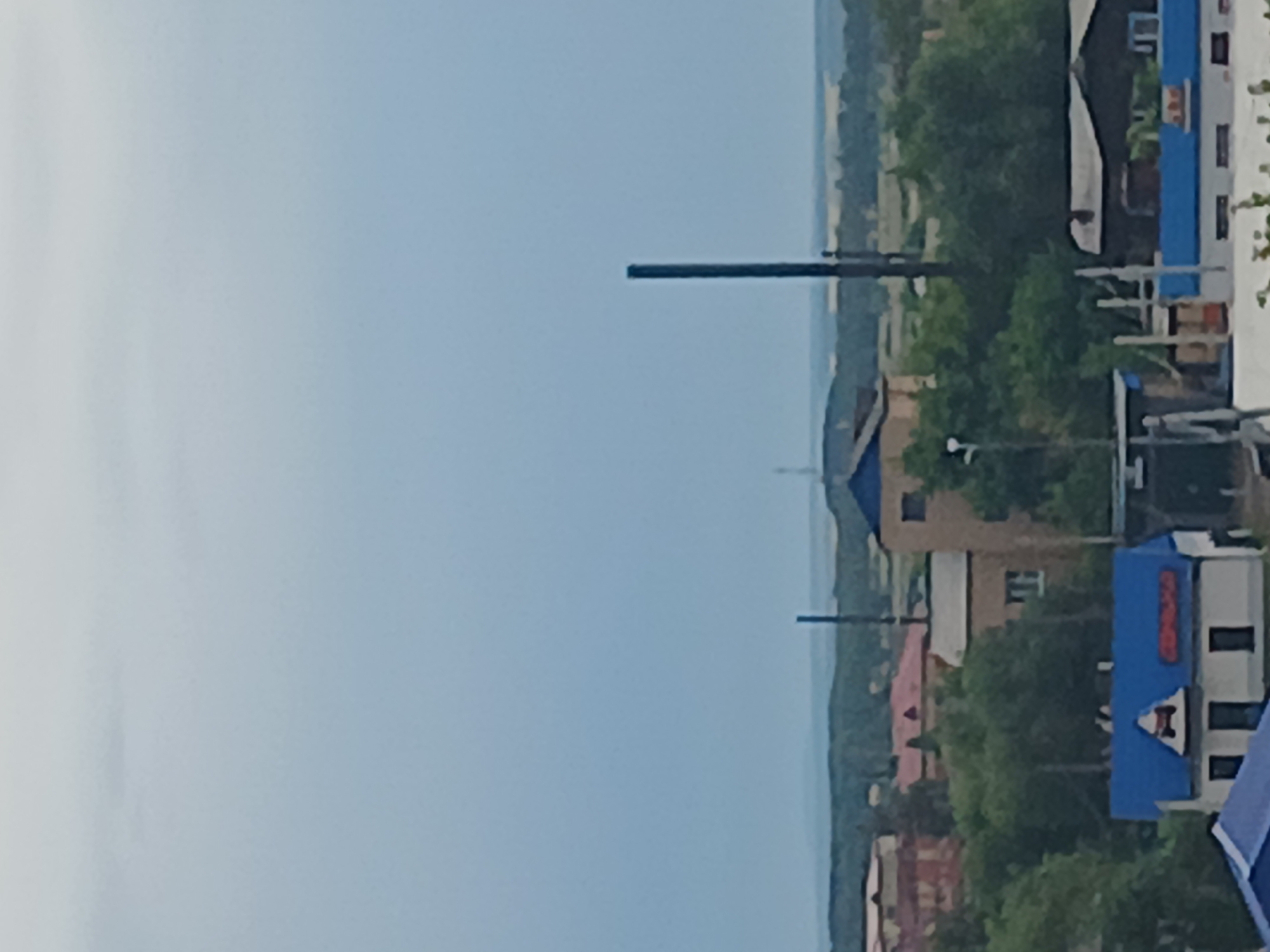 The image captures an elevated view of Makinsk, Kazakhstan, under a hazy, overcast sky. The foreground is dominated by several buildings, including one with a bright blue roof adorned with a triangular sign featuring a stylized red car and what appears to be Cyrillic lettering. Adjacent to this building, another structure also has a blue roof. Further back, more buildings with red and beige roofs are visible, interspersed with lush green trees. A tall, dark industrial chimney stands prominently in the mid-ground, with a fainter tower or structure visible on the horizon. The landscape extends to a distant, hazy horizon line, suggesting a rural or semi-urban setting on the outskirts of the city. No people are clearly discernible in the image.