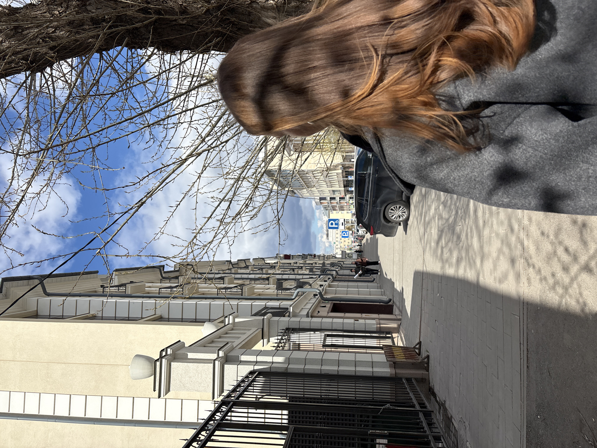 The image captures a street scene in Kazan, Russia. Buildings line the street with light-colored facades and windows. A black metal fence runs along the bottom of the image. Bare tree branches are visible in the upper left. A gray car is parked along the street, and two people are walking in the distance. A blue parking sign is visible further down the street. A person with brown hair and wearing a gray jacket stands in the foreground, partially obscuring the view. The sky is blue with scattered white clouds.