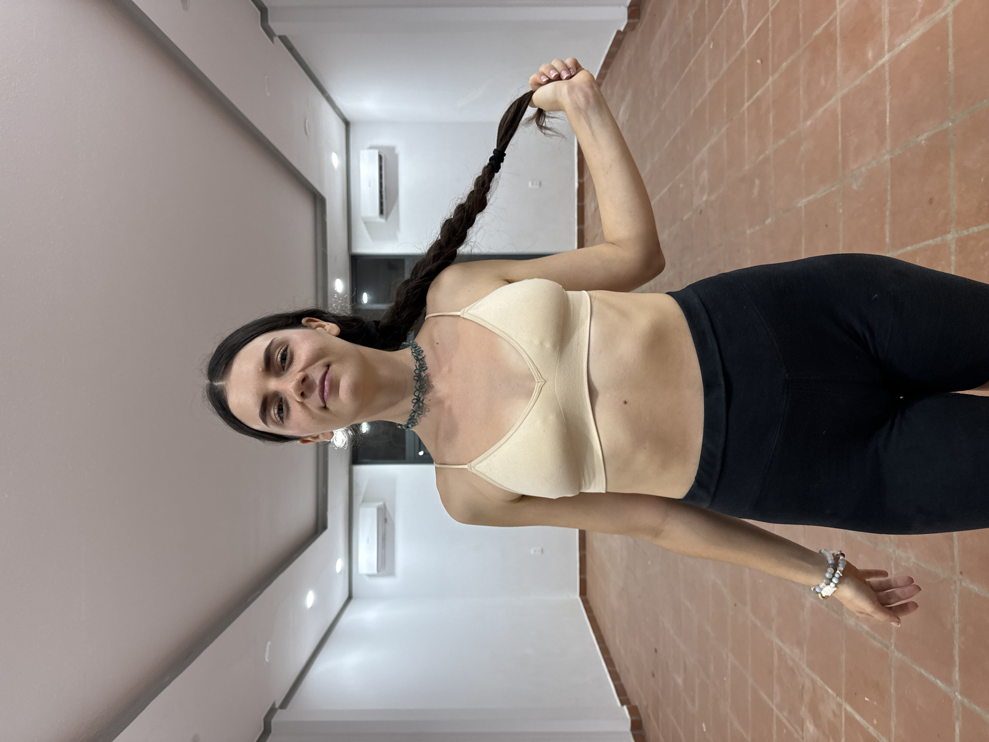 A woman with dark hair braided and held up with her right hand is in a room with a tiled floor. She is wearing a tan sports bra, black leggings, and two bracelets on her left wrist. She also has a blue beaded necklace and hanging earrings. The walls are white and a window air conditioning unit is visible. It's difficult to pinpoint the precise location in Mũi Né, Vietnam without more context.