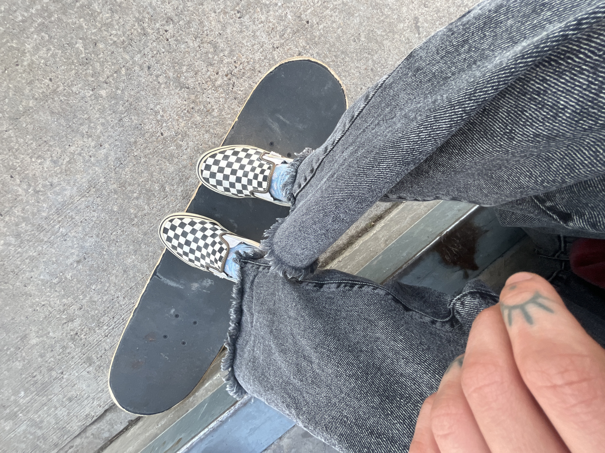 A person stands on a skateboard on concrete, wearing black and white checkered Vans shoes and grey denim pants. The pants have a frayed hem. A visible hand shows a tattoo on one of the fingers. The location appears to be an outdoor setting, possibly a skate park or urban environment, but there's no indication it is specifically Buenos Aires, Argentina.