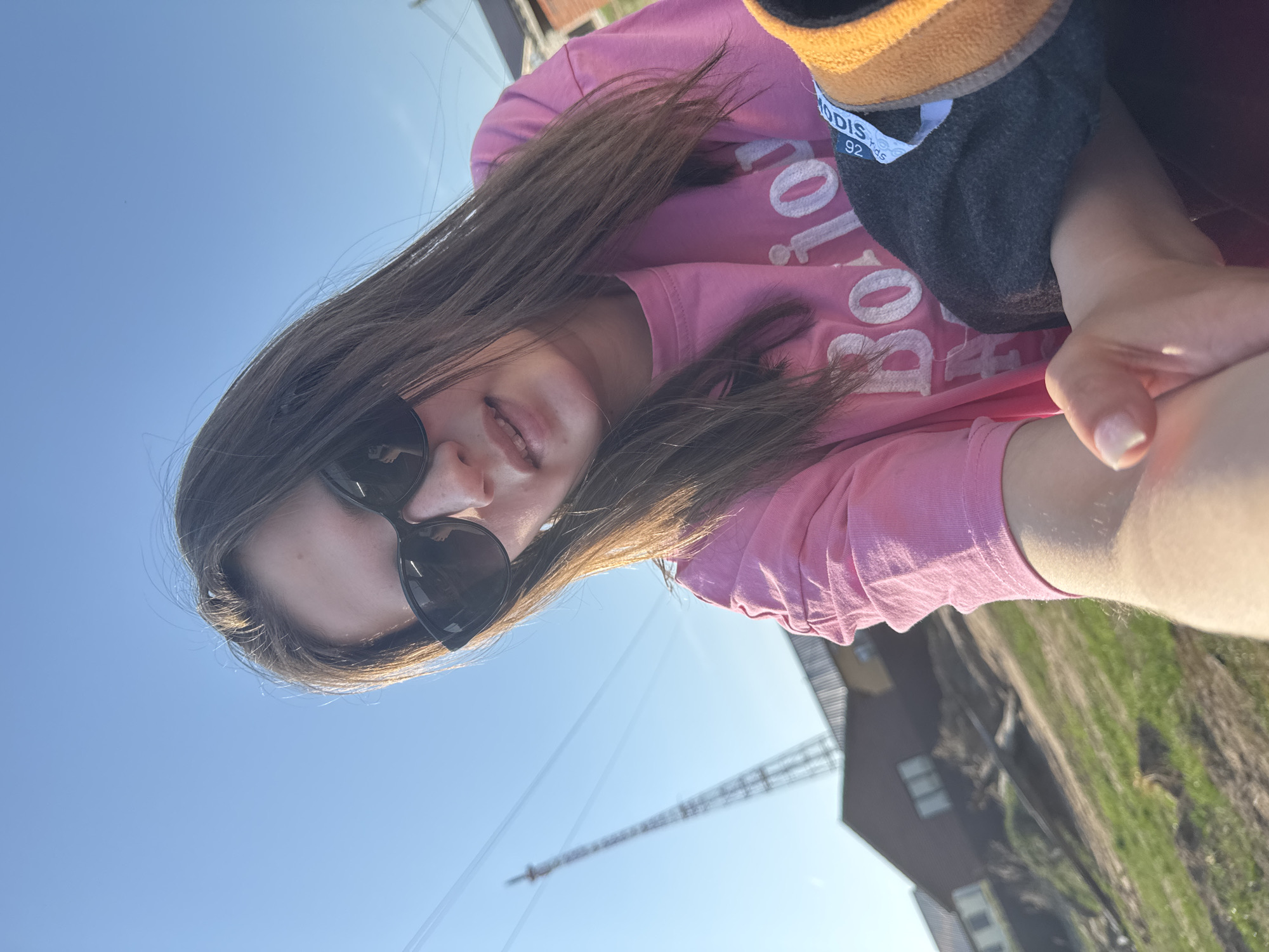 A young woman with long brown hair and sunglasses is taking a selfie outdoors on a sunny day. She is wearing a pink shirt with white lettering. She is holding a dark grey soft toy or puppet with a yellow trim. The background includes a blue sky, power lines or a crane, a building, and a grassy area. Based on the tags, this photo was taken in Chaikovsky, Russia.