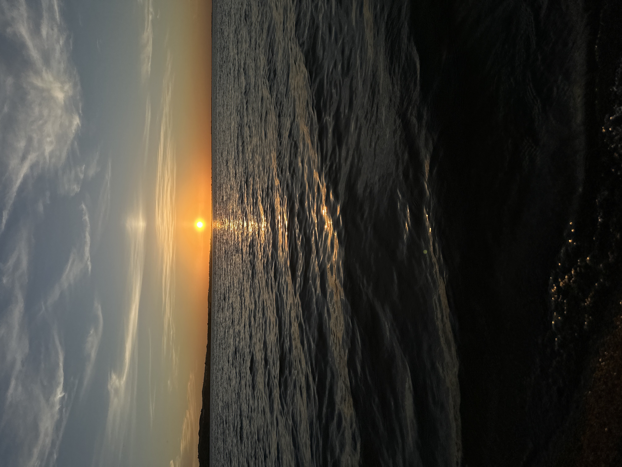 The image depicts a sunset over a body of water. The sun, a bright orange orb, is positioned near the horizon, casting a golden reflection across the rippling surface of the water. The sky above transitions from a pale orange near the horizon to a soft blue with scattered wispy clouds higher up. A dark, silhouetted landmass, possibly hills or a coastline, is visible in the far distance along the horizon. The foreground of the image is dominated by the dark, textured water, with hints of light glinting on the crests of the small waves. The overall scene is serene and natural, capturing the ambient light of dusk. This scene is not specifically identifiable as being in Kokshetau, Kazakhstan, as there are no unique landmarks or geographical features present that would definitively place it there.