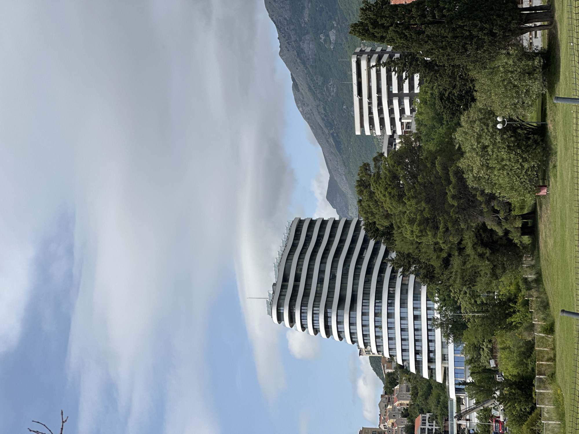 The image depicts a modern architectural scene in Bečići, Montenegro, featuring two striking, white, high-rise buildings with distinctive linear designs. The building in the foreground has a curvaceous, wave-like facade. A tree-lined park area is visible on the right, with a portion of a metal fence. Behind the buildings rises a partially cloud-covered mountain range. The sky is filled with white and gray clouds over a blue background. In the background, there are some vehicles and what appears to be a small town with buildings.