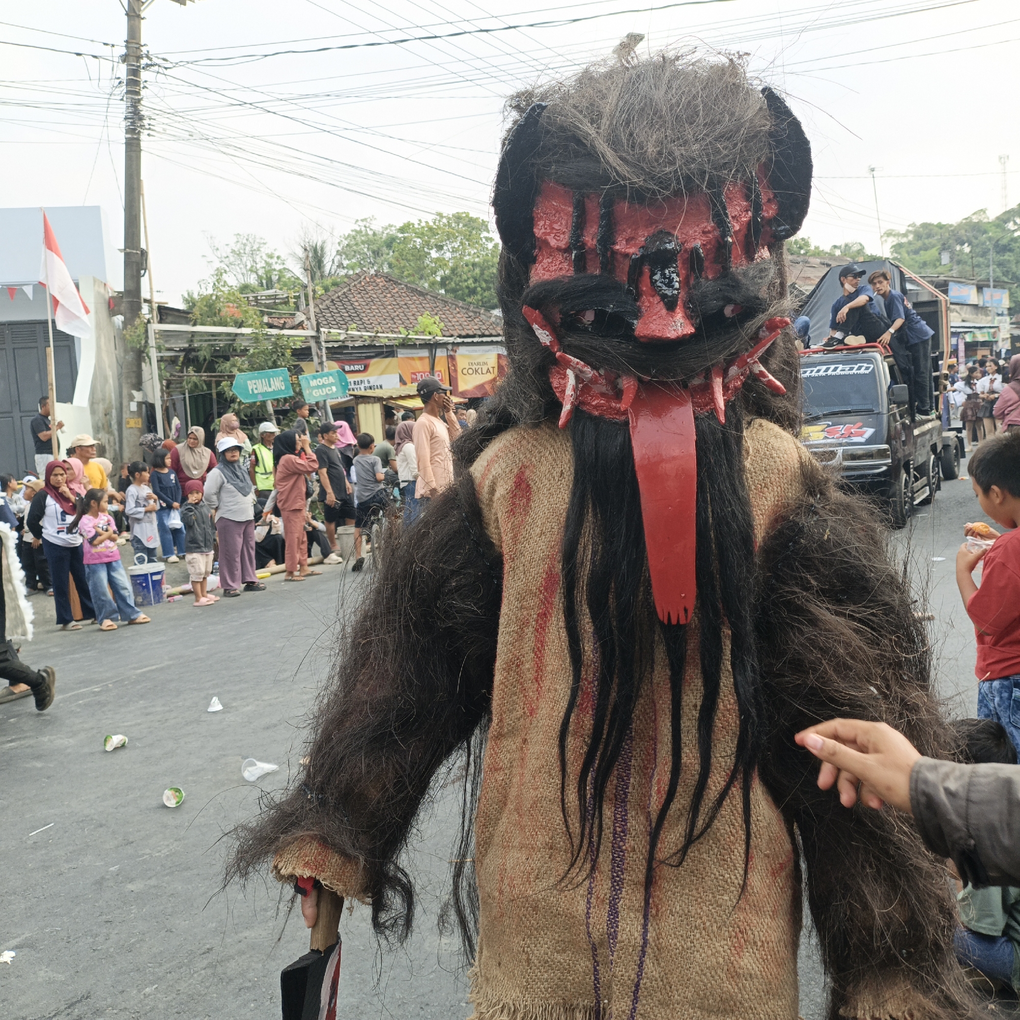 The image depicts a street scene in Randudongkal, Indonesia, possibly during a local event or parade. In the foreground, a person wearing a large, hairy costume with a red and black mask is the central focus. The mask has a grotesque appearance with exaggerated features and a long, red, forked tongue. The costume itself is made of burlap and covered in dark, shaggy hair. The person is holding a prop that resembles an axe with red markings.
A crowd of people, including men, women, and children, are visible in the background, lining the street and observing the spectacle. Some individuals are standing, while others are sitting or standing on top of a flatbed truck. The truck, a black utility vehicle, has people on its bed and appears to be part of the procession or display. Banners and signs are visible along the street, some of which are in Indonesian. One sign reads "PEMALANG" and another has text in yellow. The environment suggests a typical street in a town or city, with buildings, electrical poles, and overhead wires. The ground is paved and shows some scattered debris, including discarded cups. The overall atmosphere appears to be one of a public gathering or celebration.