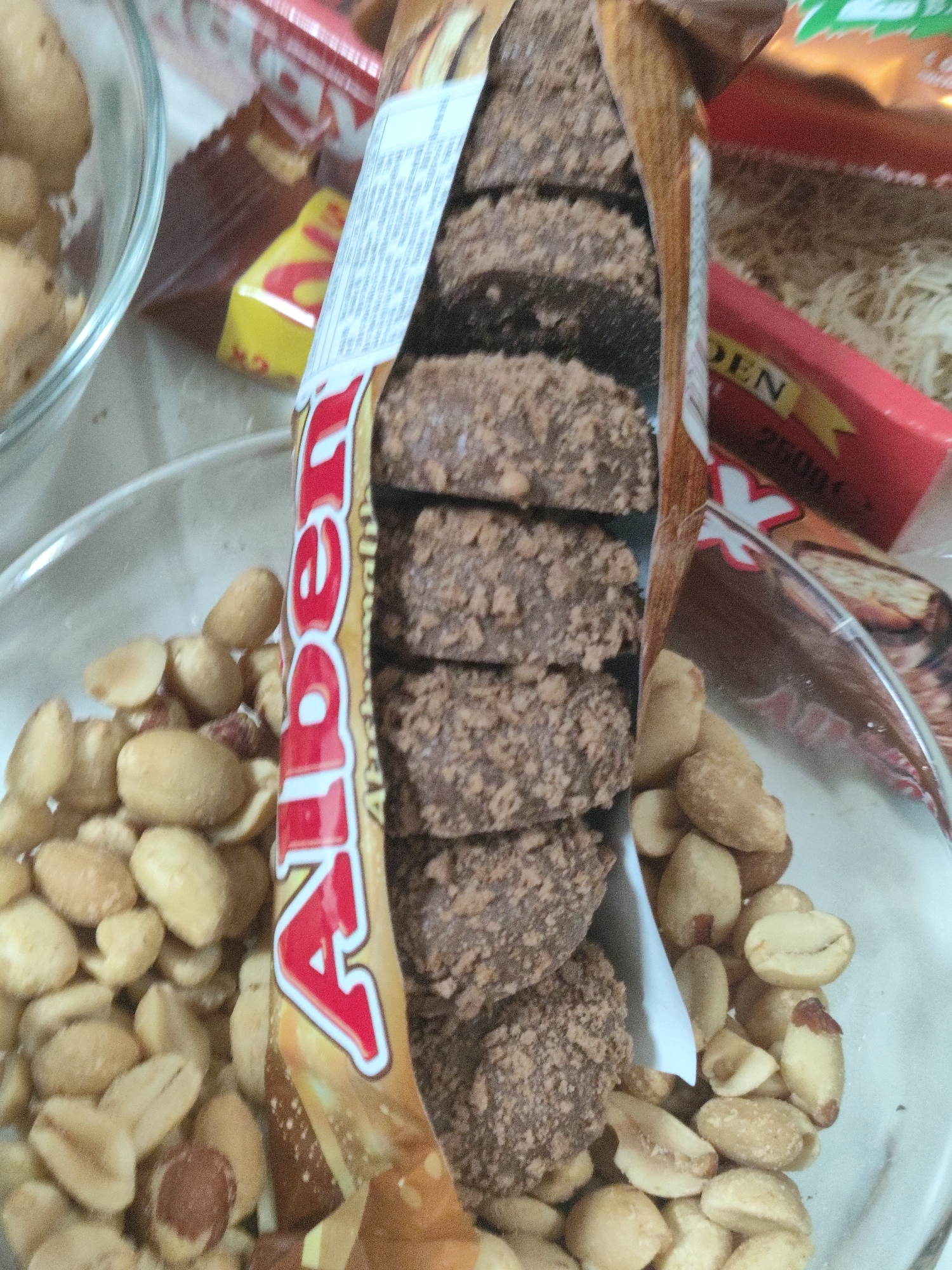 The image displays a close-up view of a snack selection, prominently featuring a package of "Alben" chocolate biscuits, a glass bowl filled with peanuts, and other snack wrappers in the background. The Alben package is partially opened, revealing a stack of chocolate-covered biscuits. The peanuts are shelled and appear to be a mix of whole and halved nuts. The overall setting suggests a casual, indoor environment, likely a home or a small gathering. There is no indication of a specific location in Asan-si, South Korea, within the image itself.