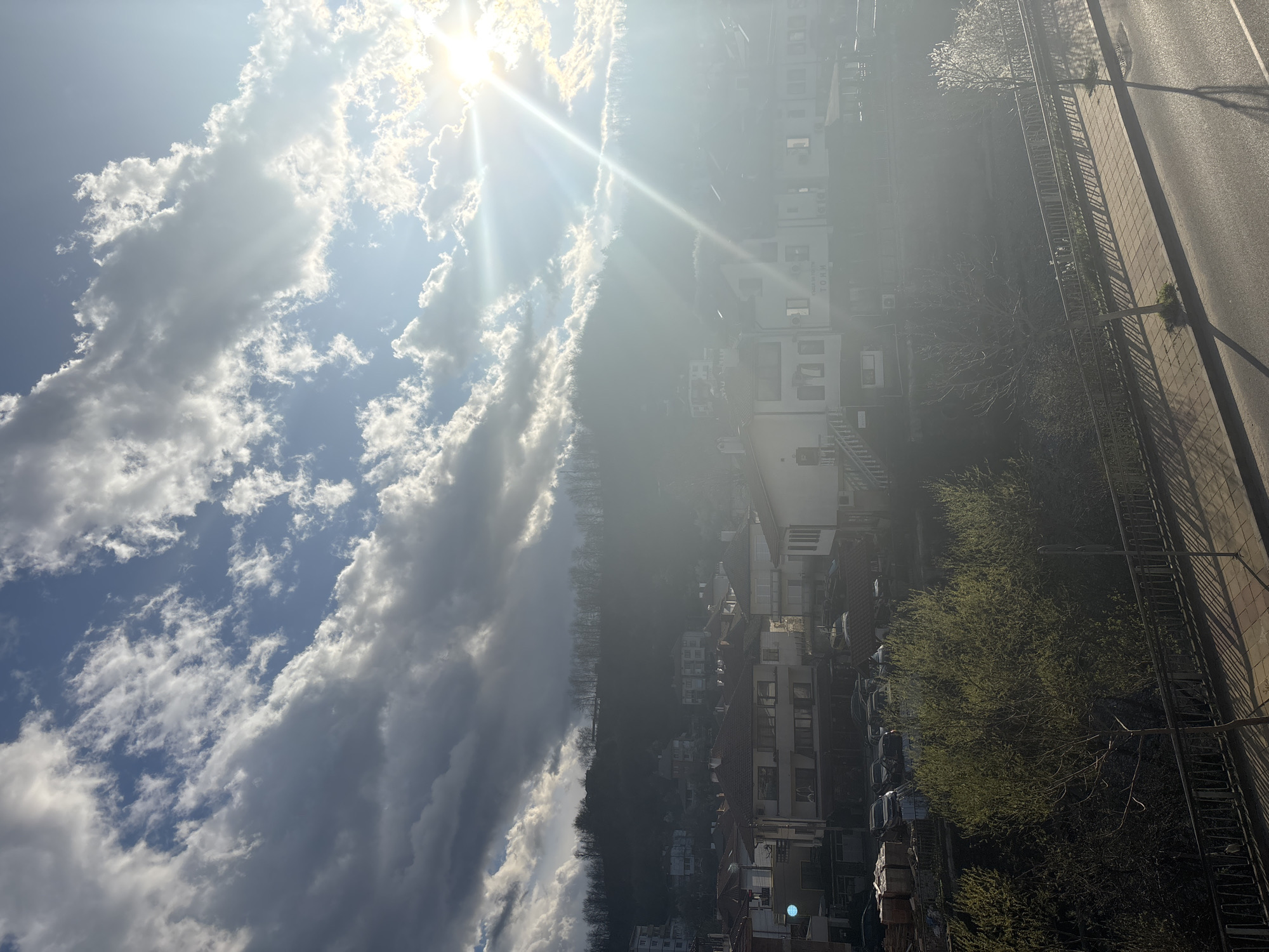The image depicts a cityscape in Zlatograd, Bulgaria, under a bright, sunlit sky with scattered clouds. The sun is positioned high in the sky, casting strong rays. The foreground shows buildings, roads, trees, and vehicles. The overall perspective is from a high vantage point, potentially from a building or elevated location. The scene exhibits natural sunlight and atmospheric conditions.