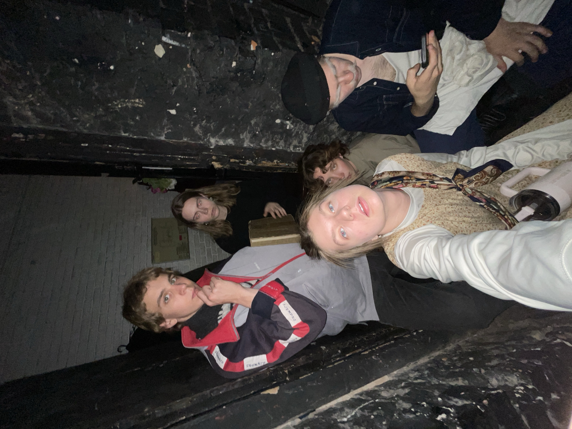 Six individuals are positioned in what appears to be an enclosed, possibly derelict space with worn walls. The space is cramped, as evidenced by the proximity of the subjects to one another and the limited visibility of the environment. The lighting is dim, which contributes to a gritty atmosphere. The individuals seem to be posing for the camera. One individual is holding a mobile phone. No indication of city/location is visible.